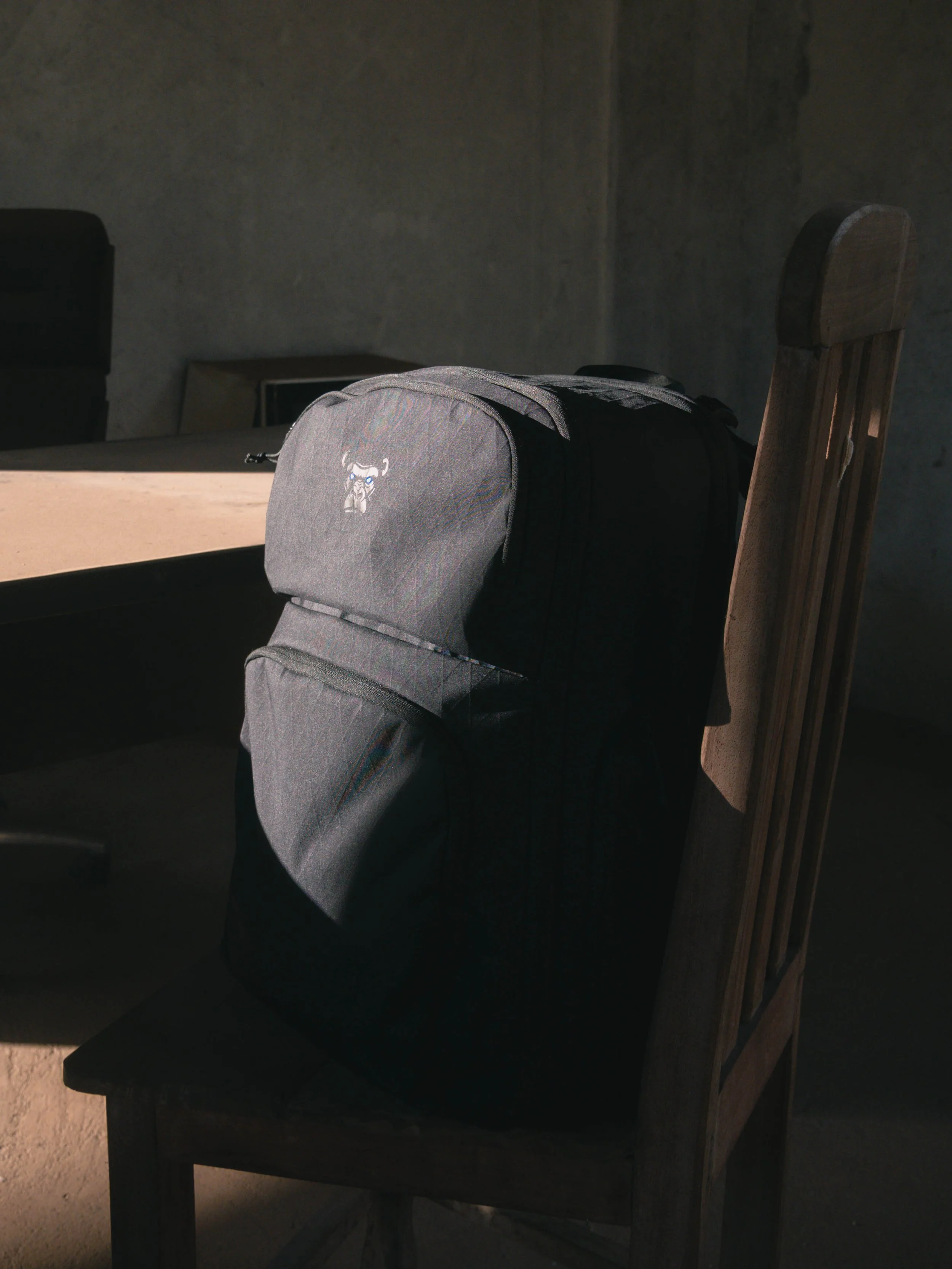 A wooden table with a gray backpack on top, and a wooden chair next to it. The room has a gray wall in the background.