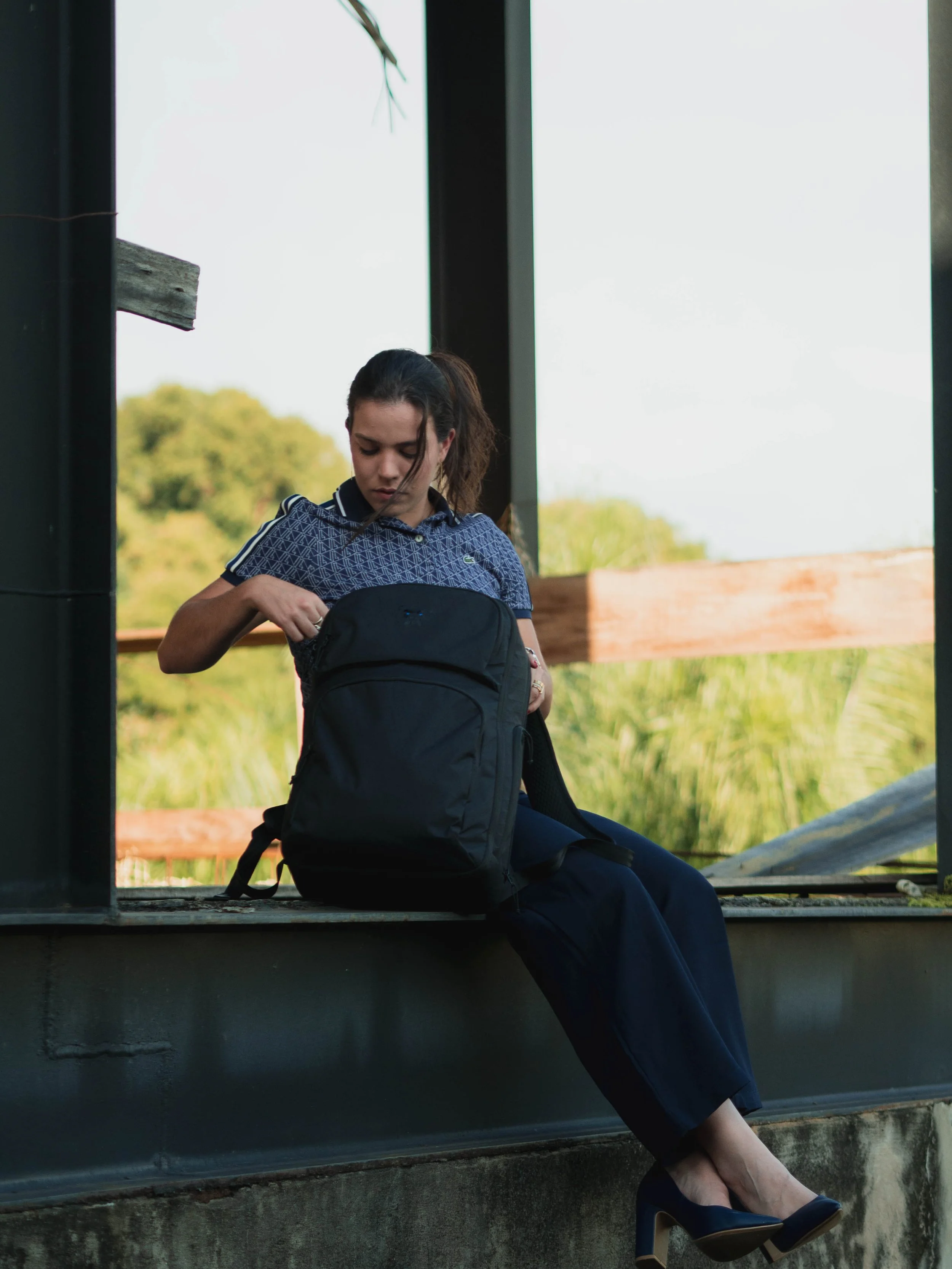 A young woman in a blue patterned shirt and dark pants sits on a low ledge, looking into her backpack. She is wearing dark high heels, and her hair is tied back. The background features a rural landscape with greenery and trees, with a partly cloudy sky.