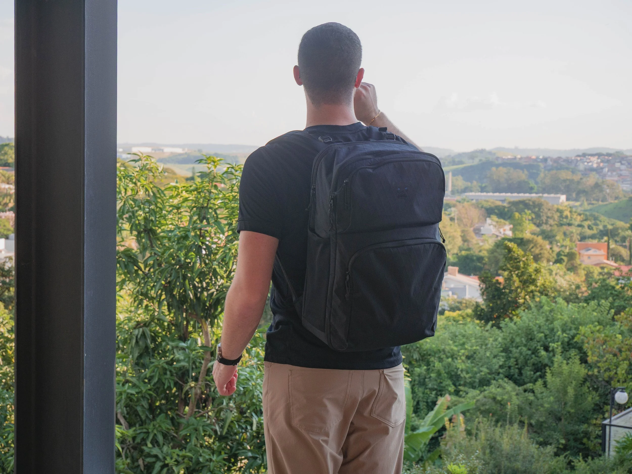 Back view of a man with short dark hair wearing a black t-shirt, khaki pants, and a black backpack, standing outdoors on a balcony or terrace overlooking a lush green landscape with buildings in the distance in daytime.
