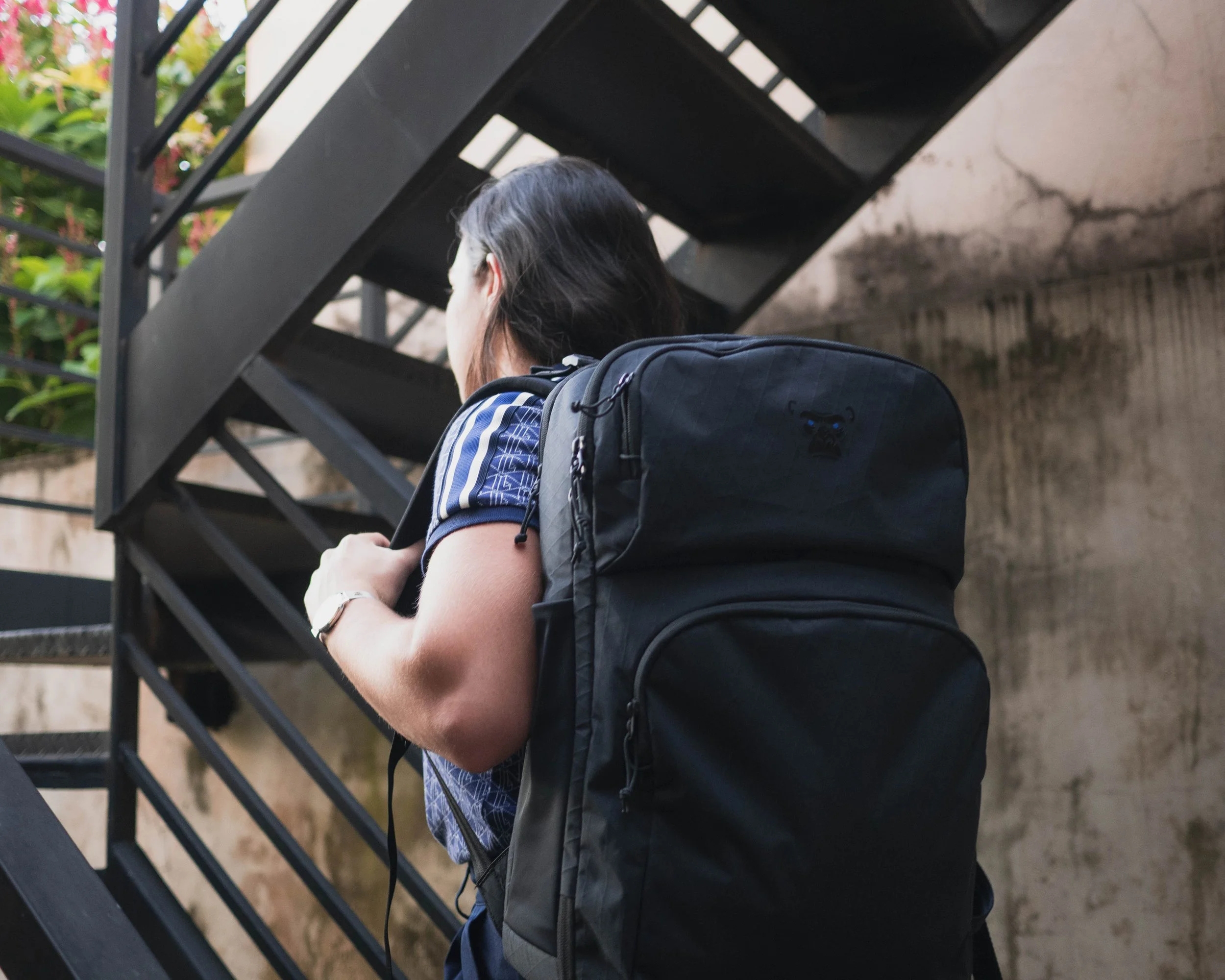 A person with long dark hair, wearing a short-sleeved shirt with blue and white patterns, is sitting on stairs with their arms crossed. They have a black backpack on their back and are looking downward. The stairs are black metal with diagonal railing bars, and there are green plants with pink flowers visible in the background.