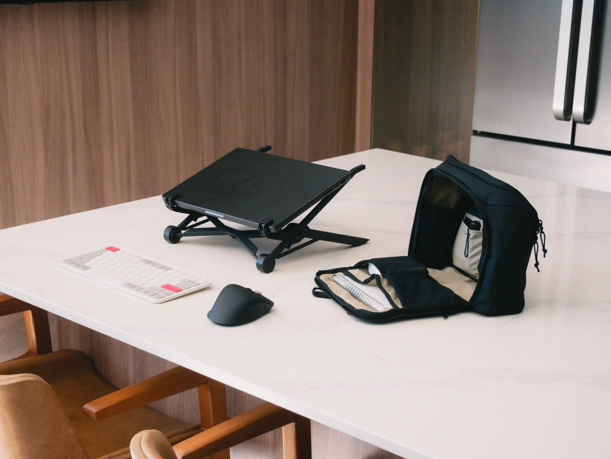 A white table with a black laptop stand, a white keyboard, a black mouse, and a black backpack on top. The table is in a room with wood-paneled walls and a refrigerator in the background.