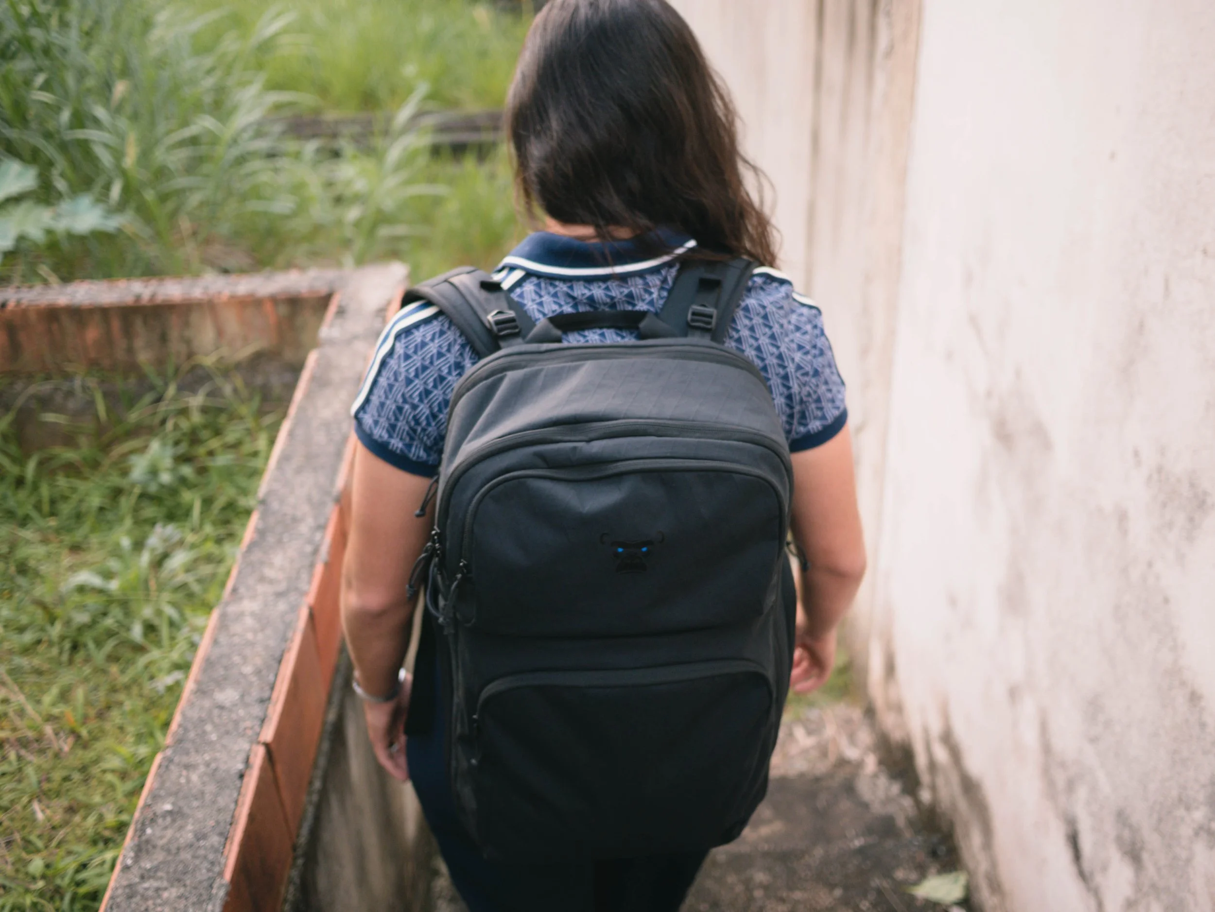 A girl with long dark hair wearing a blue patterned shirt and black backpack walking along a narrow outdoor path next to a white wall and some green plants.