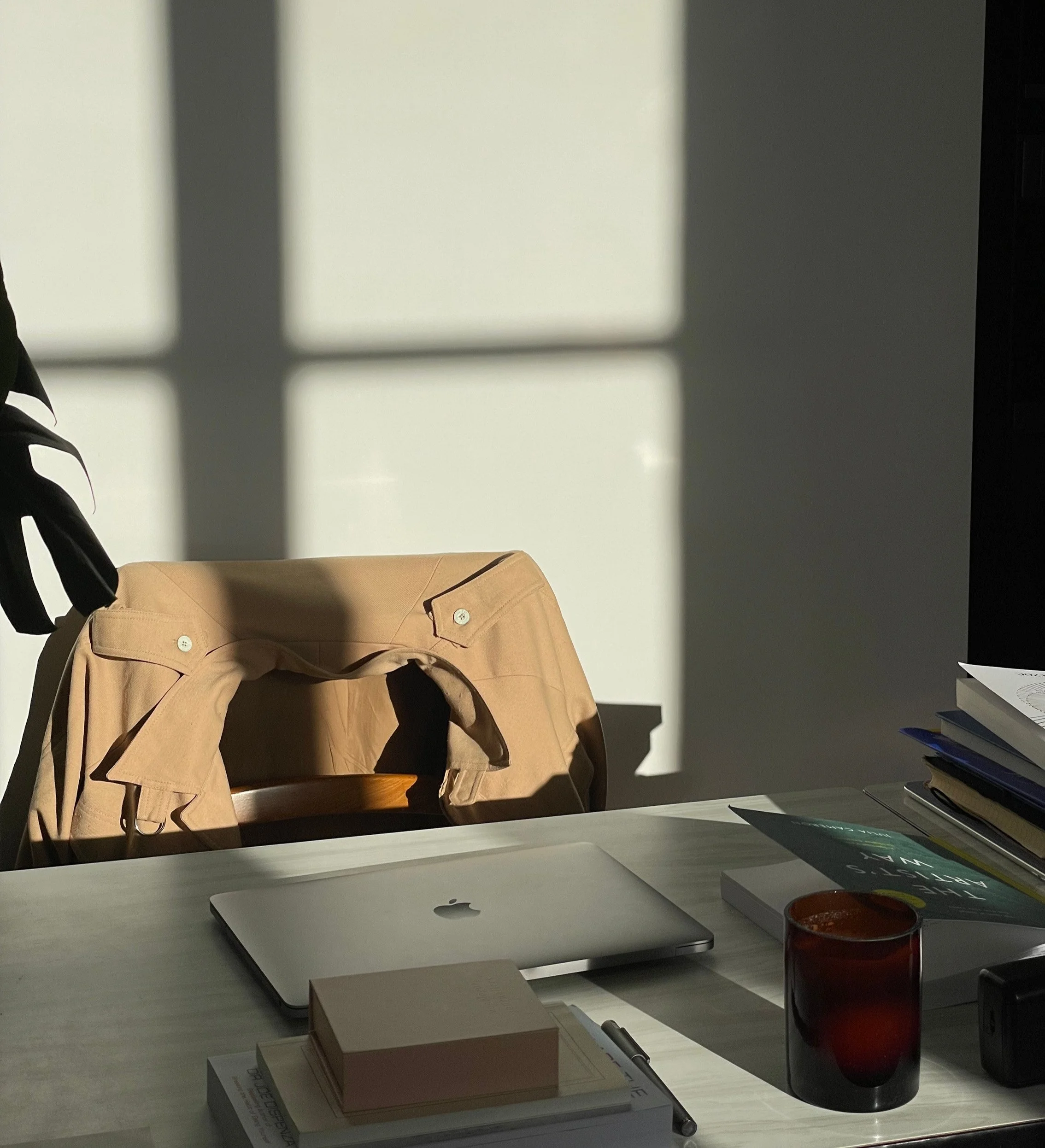 A trench coat draped over a chair in a sunlit workspace with a closed laptop, books, and a candle on the desk, representing the focused diagnostic work of The Diagnosis advisory engagement from True House Advisory.