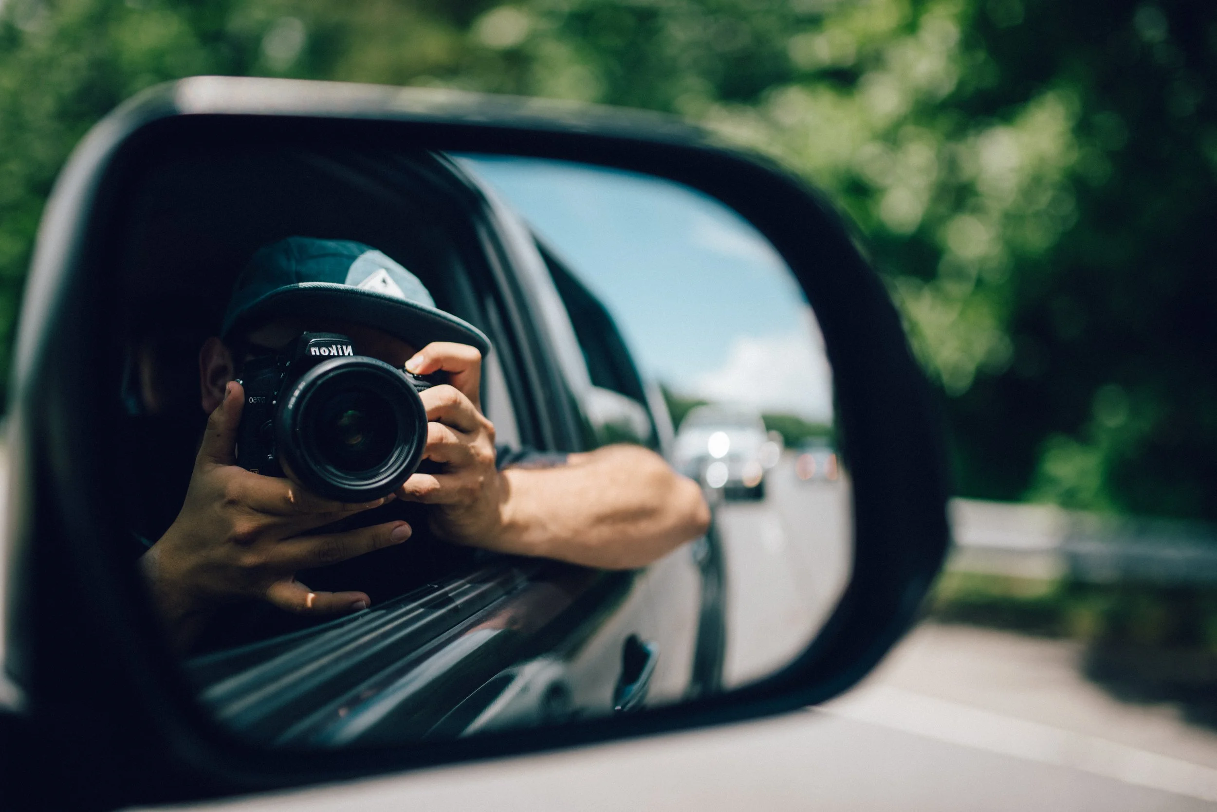 Person taking a photo with a Nikon camera while looking into a car's side mirror, with green trees in the background.