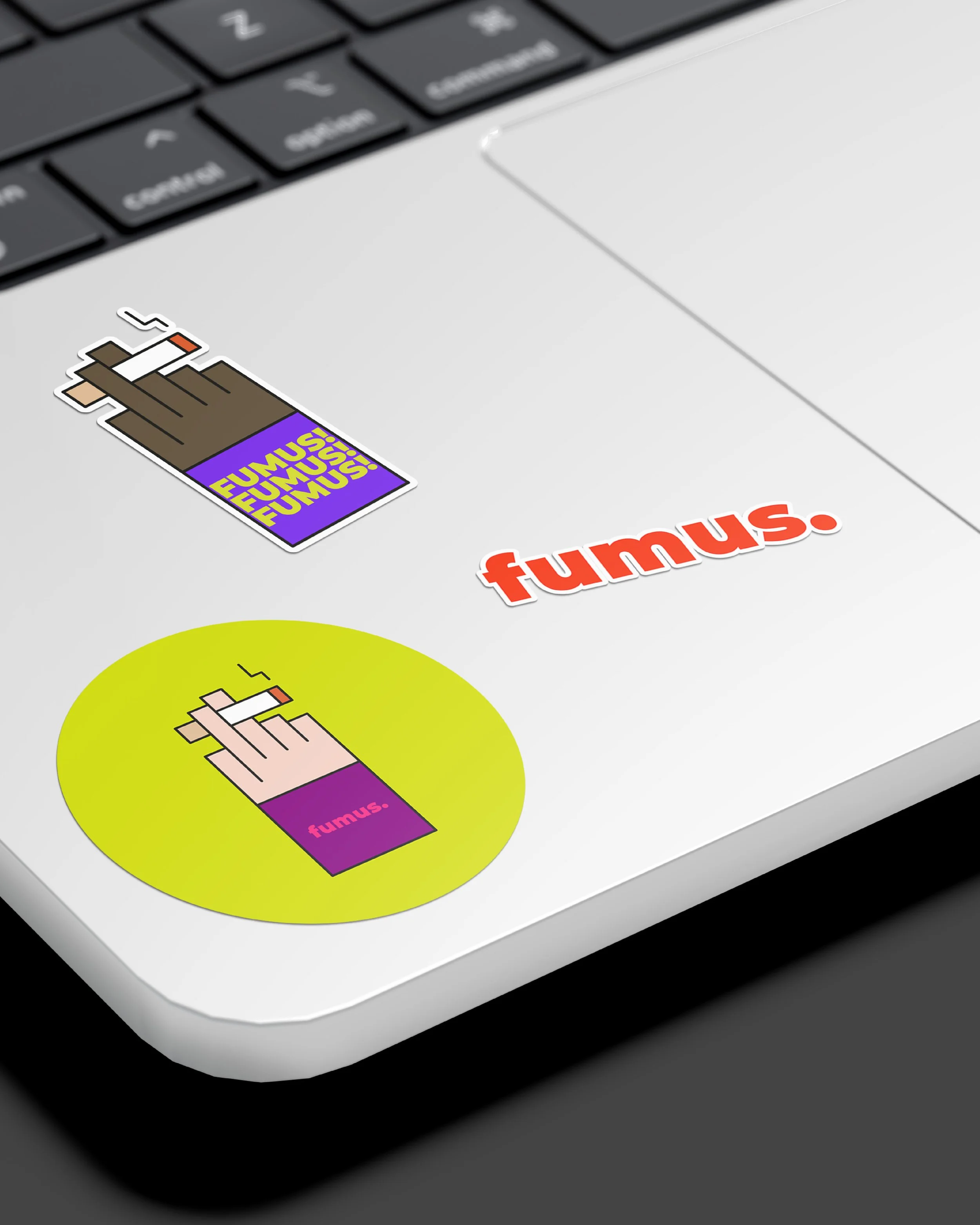 Pegatina con un dibujo de un cigarro que emite humo y la palabra 'FUMUS' en letras amarillas sobre fondo morado, colocada en una superficie de color gris claro.
