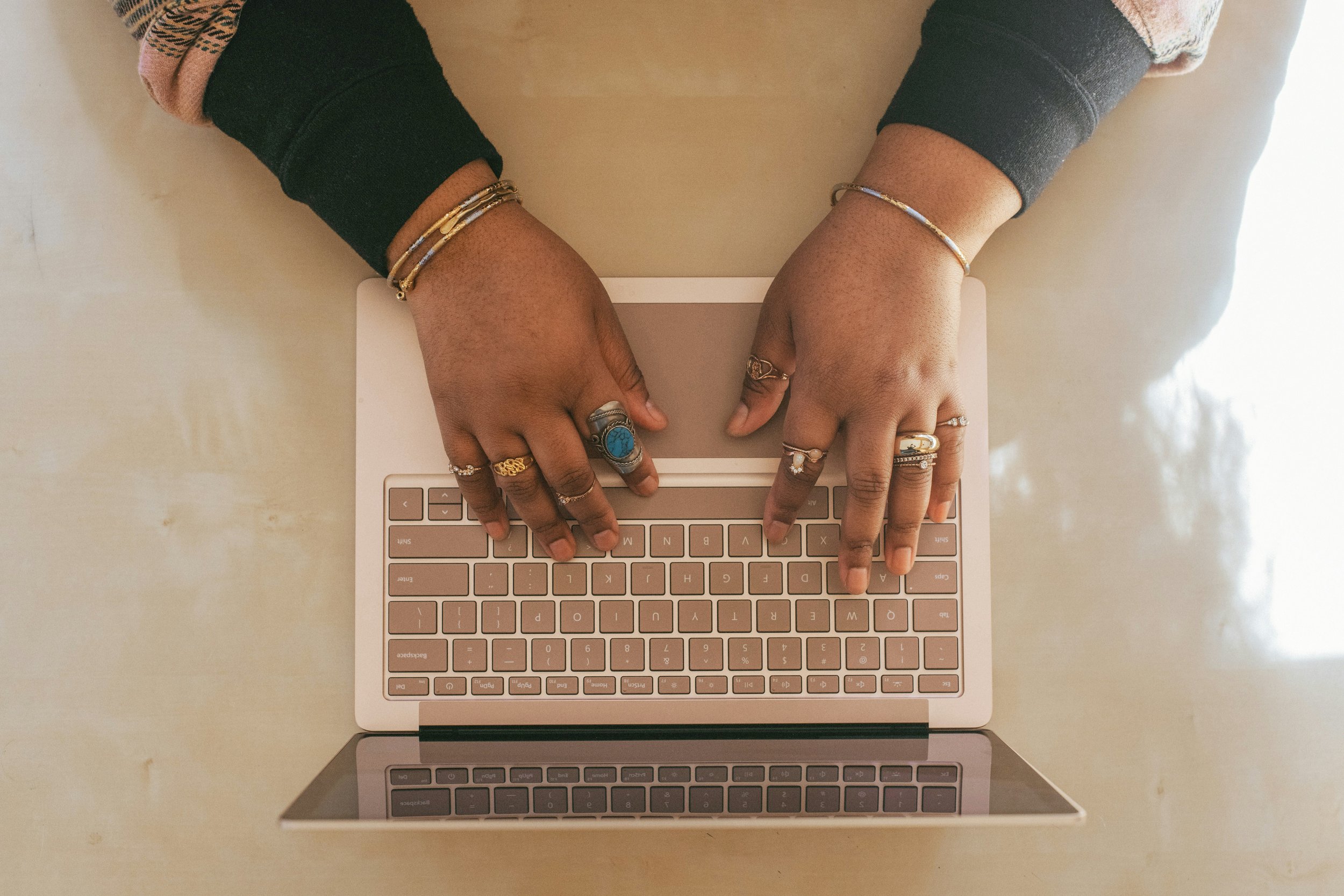 Two hands with jewelry typing on a laptop keyboard seen from above on a white surface.