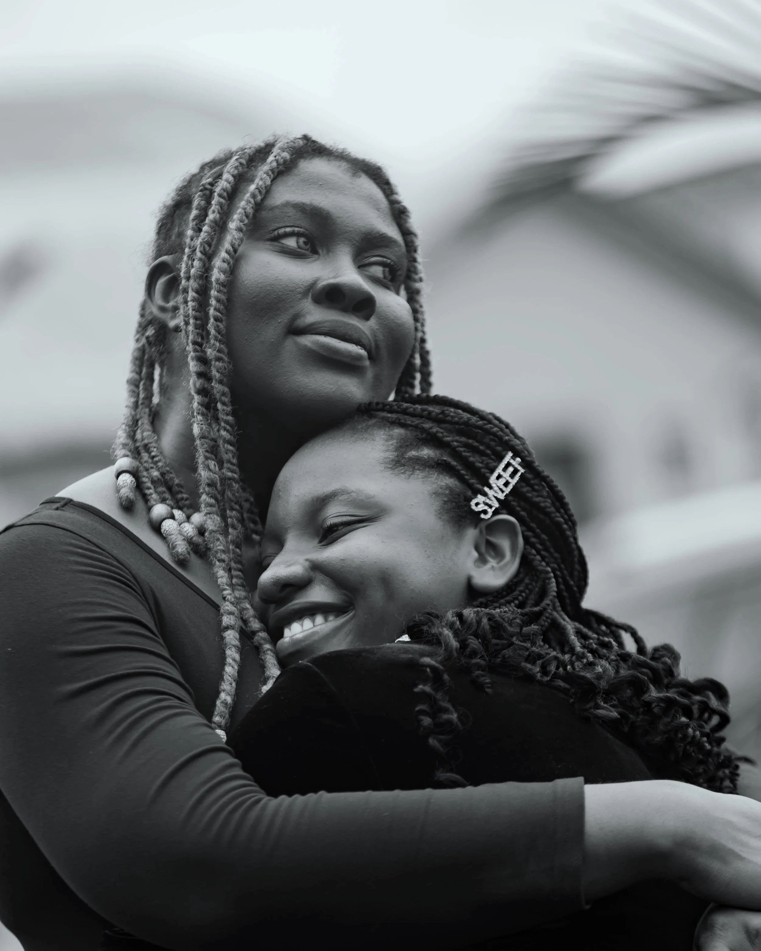 A black and white photo of two women hugging, one woman with braided hair and the other with dreadlocks, smiling.