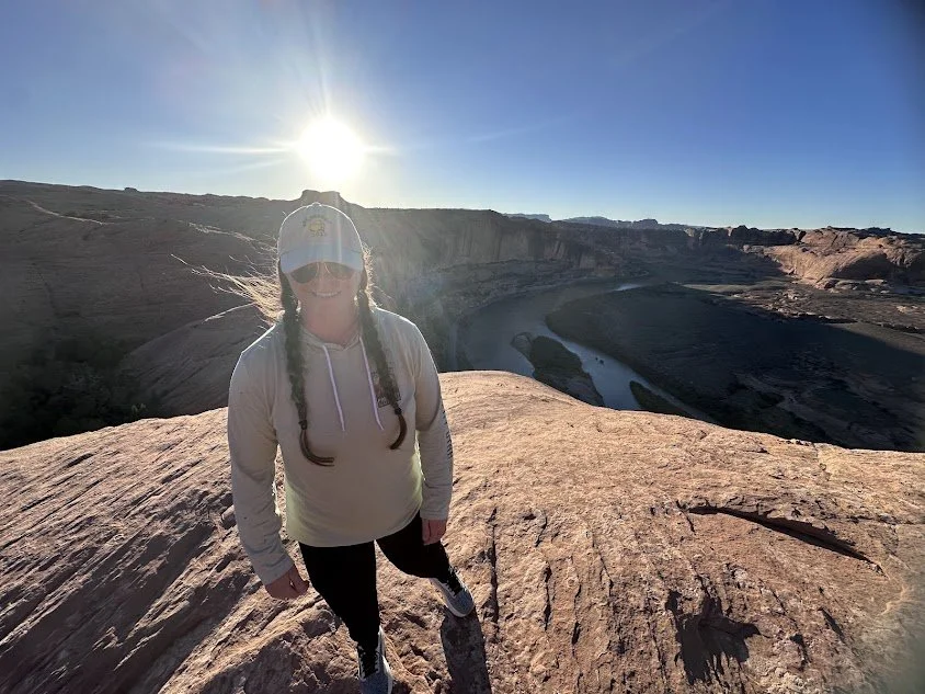 Woman with braids, wearing a hat, sunglasses, and a hoodie, standing on a rock formation overlooking a canyon and river during sunset.