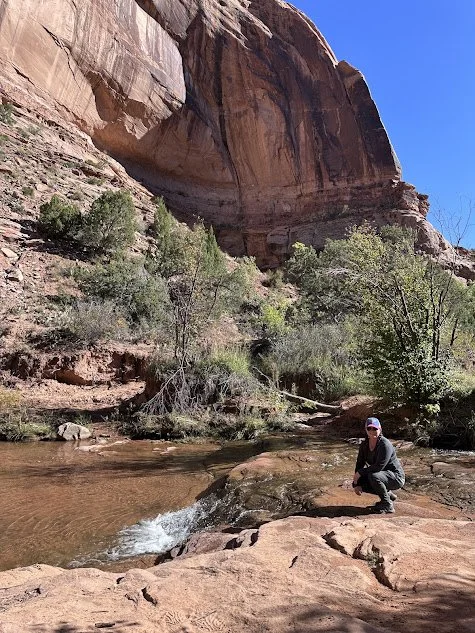 A person crouching on a large rock near a small stream in a desert canyon, with red sandstone cliffs and sparse green trees under a clear blue sky.