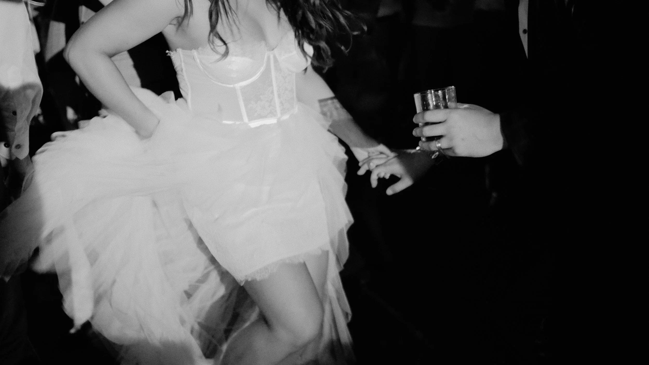 A woman wearing a white dress with lace bodice, hand at her waist, is being handed a shot glass by a person in a dark suit, with a ring on their finger.