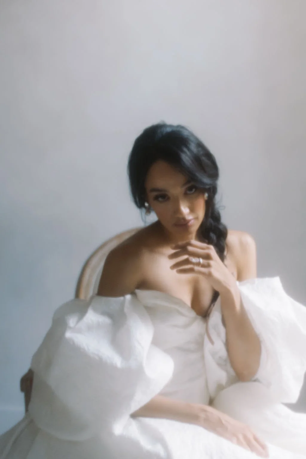 A woman with dark hair styled in loose waves, dressed in an off-shoulder white dress, sitting on a chair against a plain background, looking directly at the camera with a relaxed pose.
