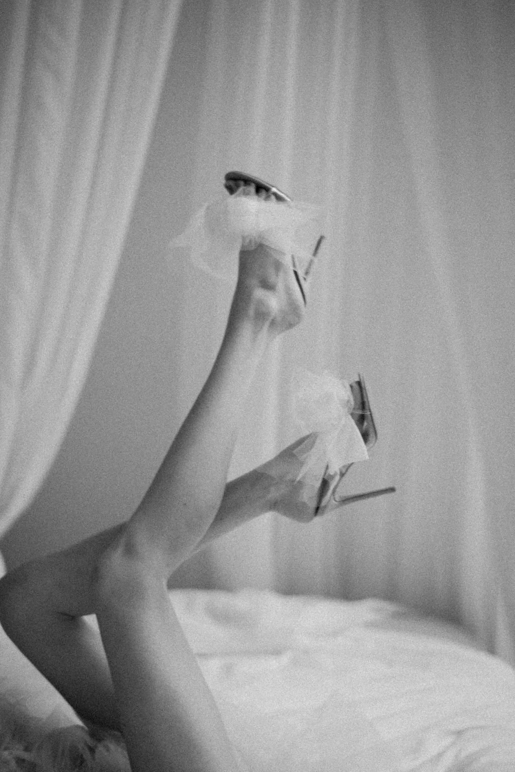 A person's legs and feet in high heels resting on a bed, with a sheer piece of fabric draped over the legs, and a curtain in the background.