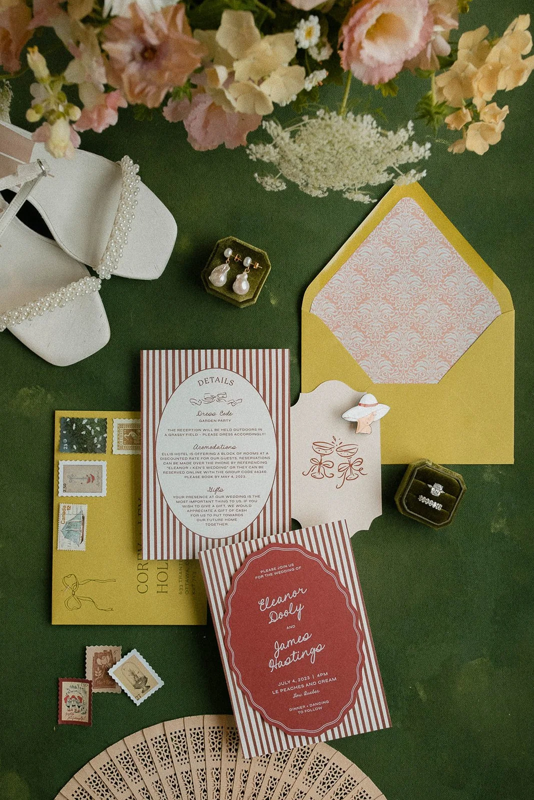 Wedding invitation cards, jewelry, postage stamps, and a decorative purse on a green surface, with pink and white flowers above.