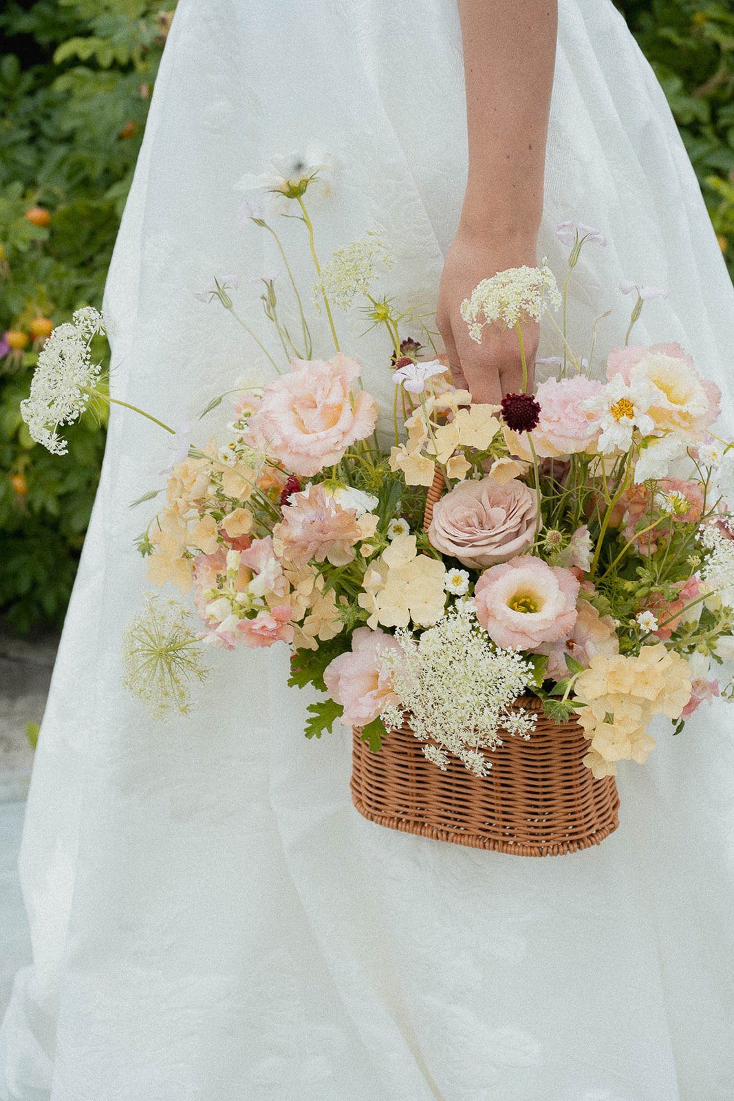 A woman in a white wedding dress holding a wicker basket filled with pastel-colored flowers, including roses, peonies, and daisies, with greenery in the background.