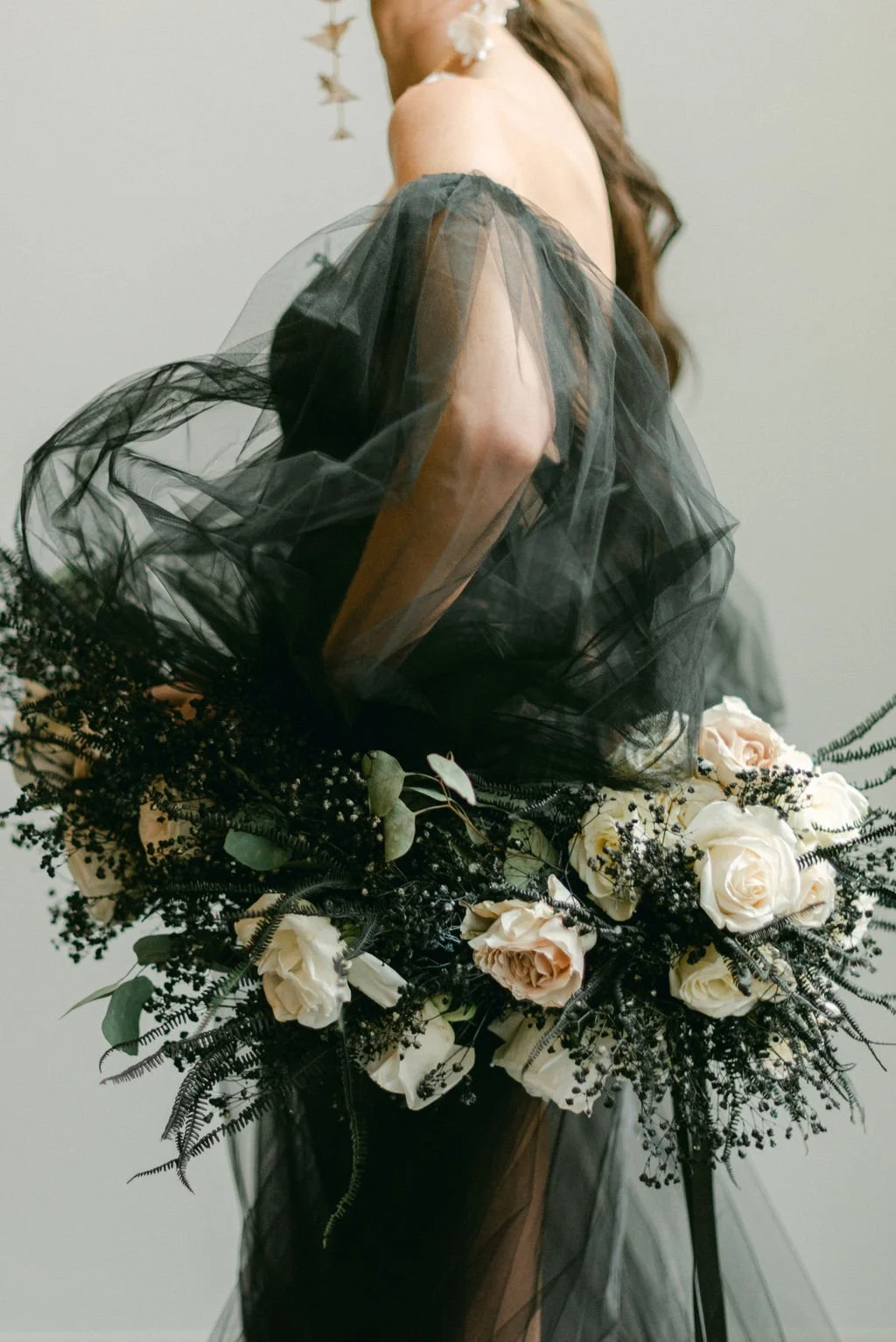 A women wearing black sheer and tulle dress, holding a large bouquet of white and blush roses with greenery and black accents.
