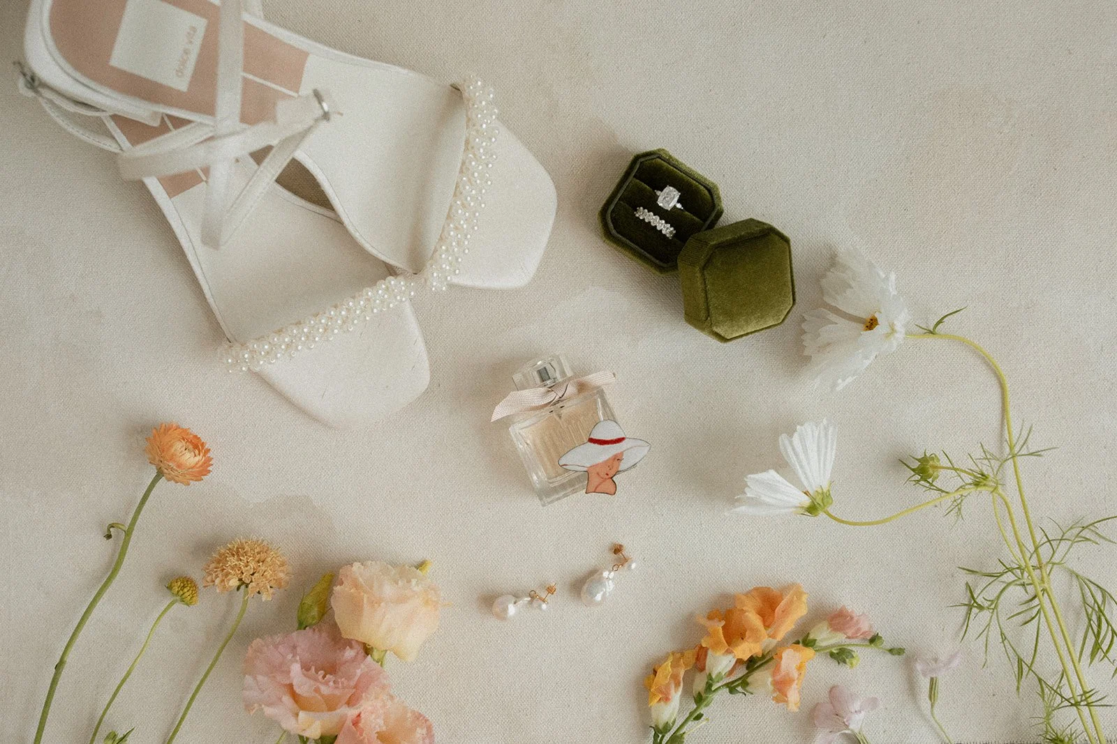 A collection of jewelry, a small perfume bottle, and flowers arranged on a cream-colored surface. The jewelry includes rings and earrings, with a white handbag decorated with pearls.