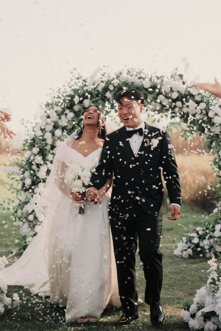 A newlywed couple walking outdoors under a floral arch, celebrating their wedding with confetti, both smiling and holding hands.