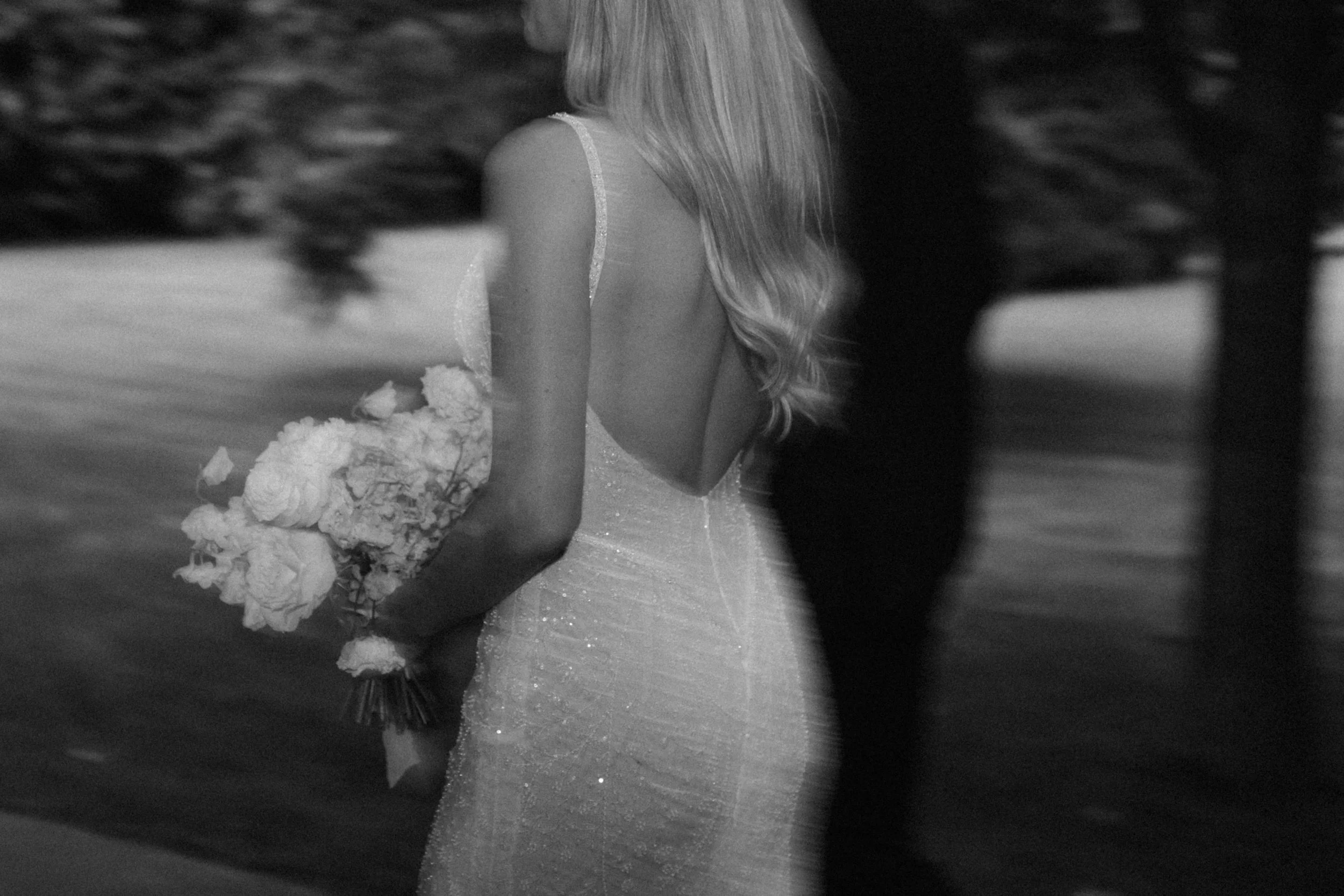 A black and white photograph of a woman in a wedding dress holding a bouquet of flowers, viewed from behind with long, flowing hair.