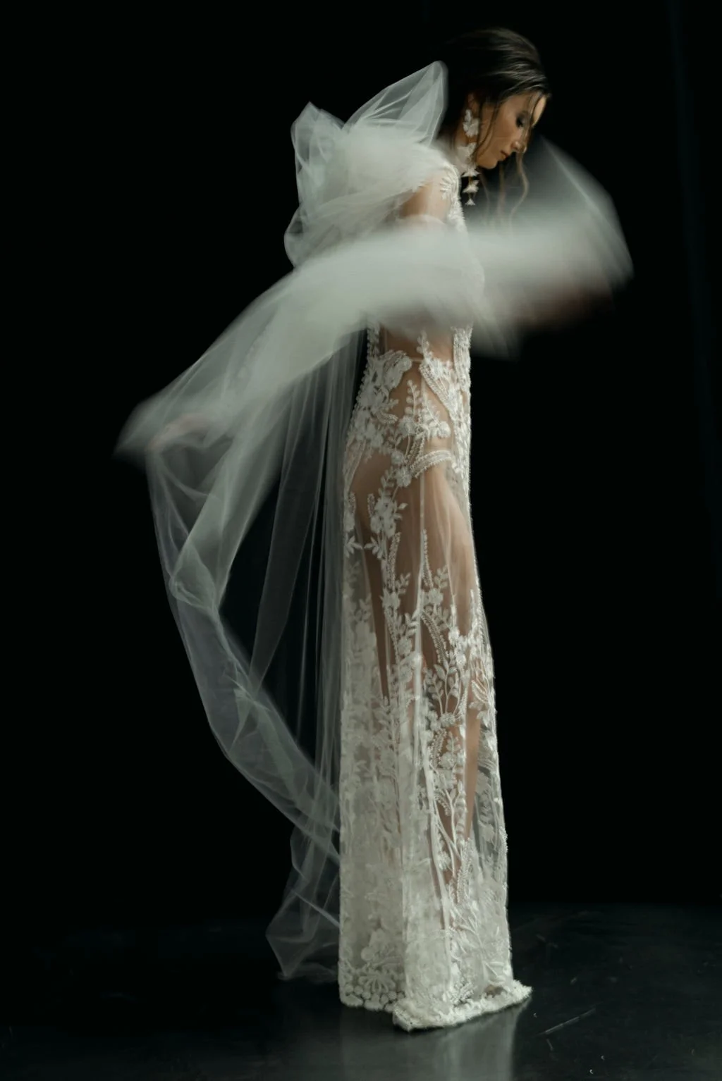 A woman in an intricate lace wedding dress with sheer panels, wearing white earrings and a veil, is adjusting her dress in a studio with a black background.