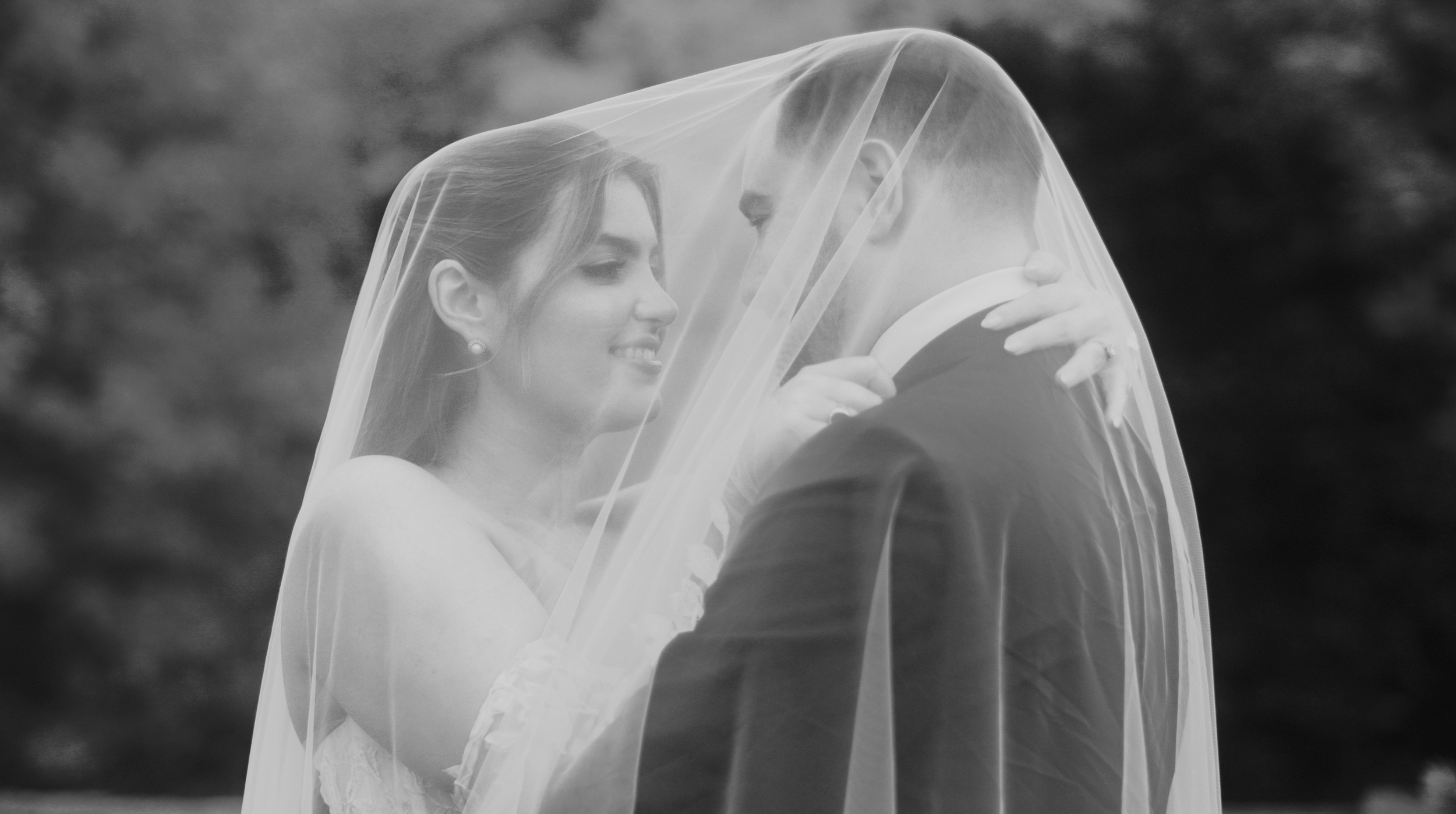 In this black and white photo, a bride and groom are close, under a sheer wedding veil. The bride is smiling and looking into the groom's eyes, while the groom gently holds the veil and looks at her. They are outdoors with blurred natural background.
