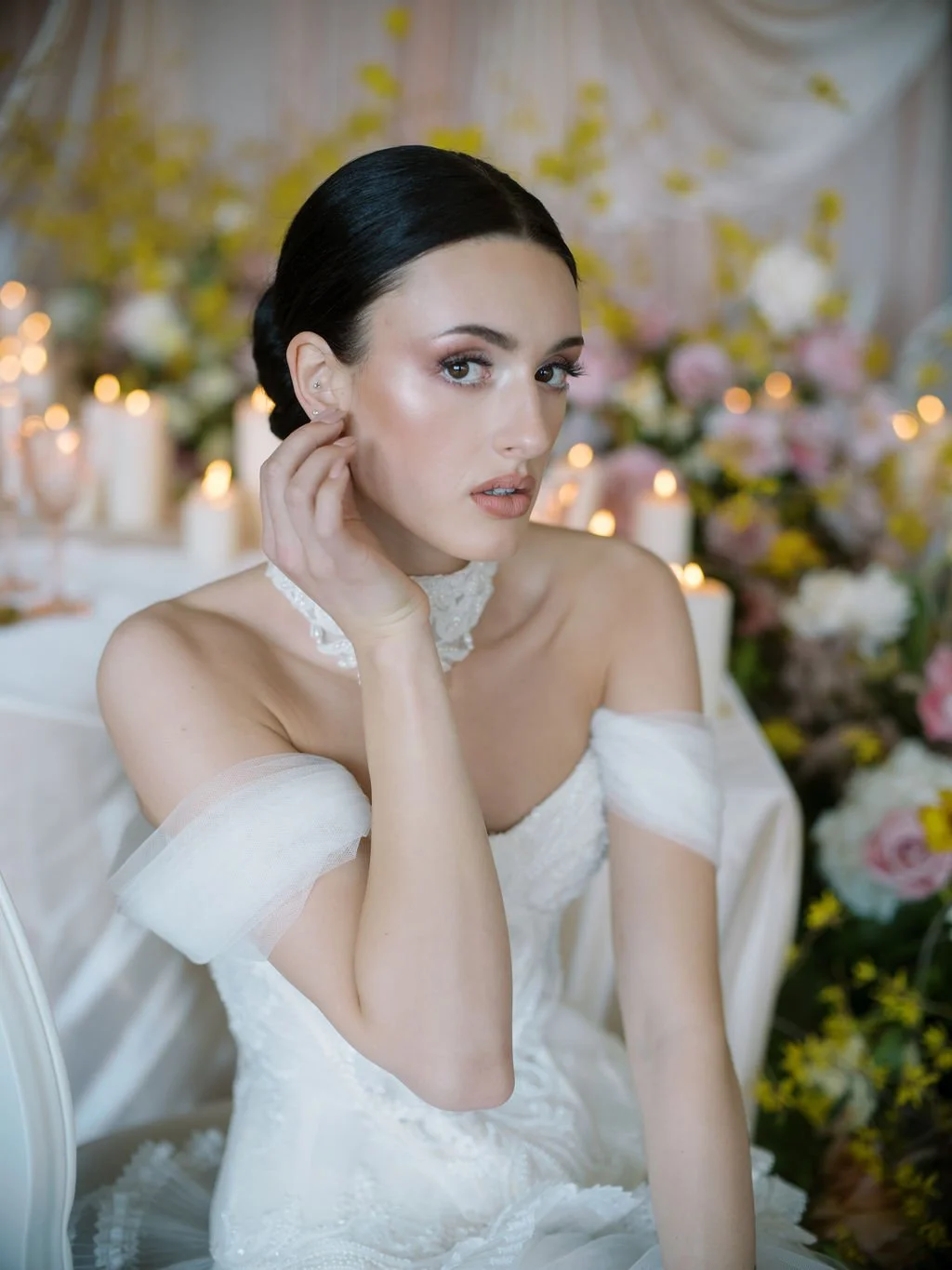 A woman with dark hair in an elegant white off-shoulder dress and lace choker, touching her face, in a decorated setting with pink, white, and yellow flowers, and lit candles.