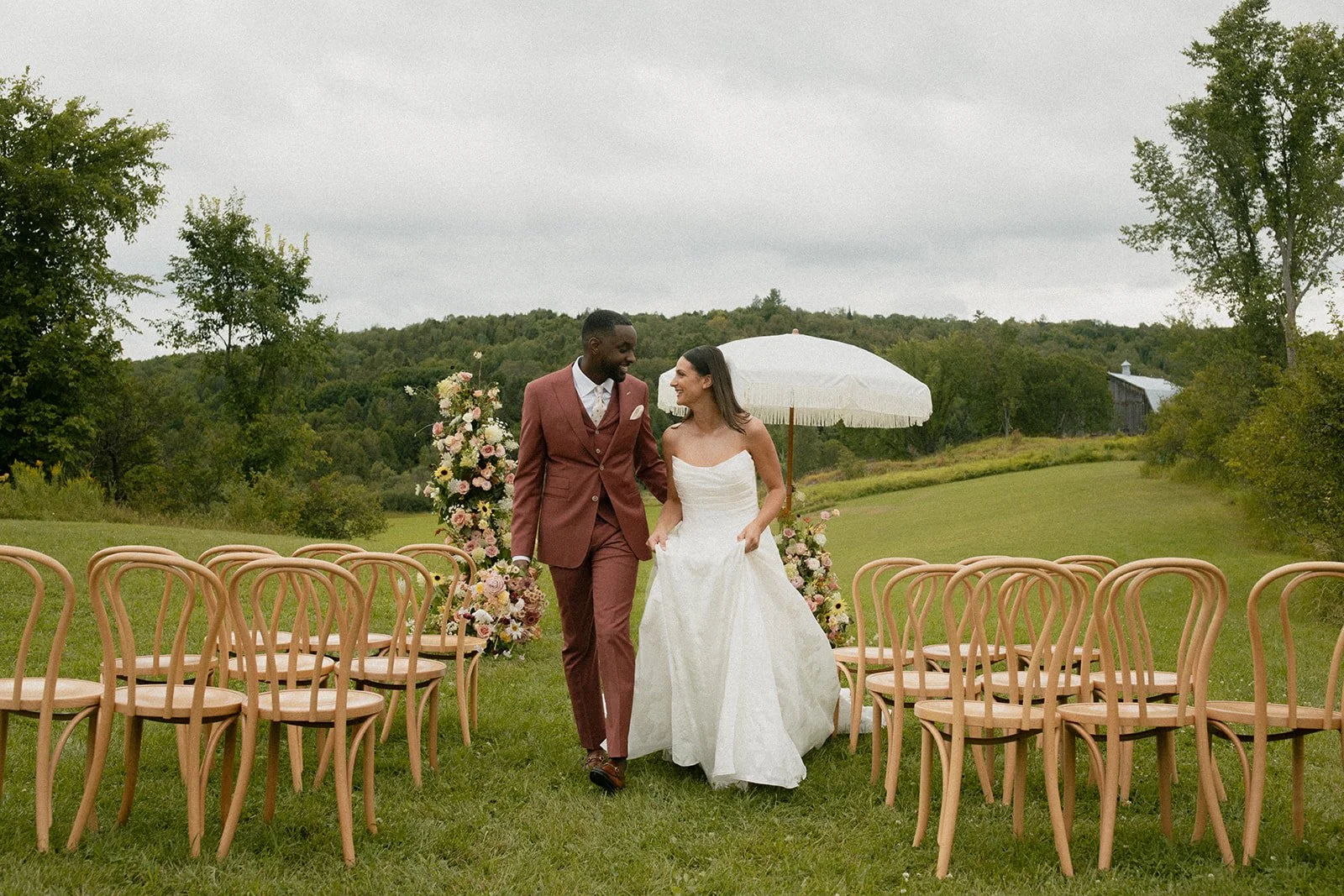 A newlywed couple walking down the aisle outdoors on a grassy field with rows of chairs, floral arrangements, and an umbrella, surrounded by trees and hills.