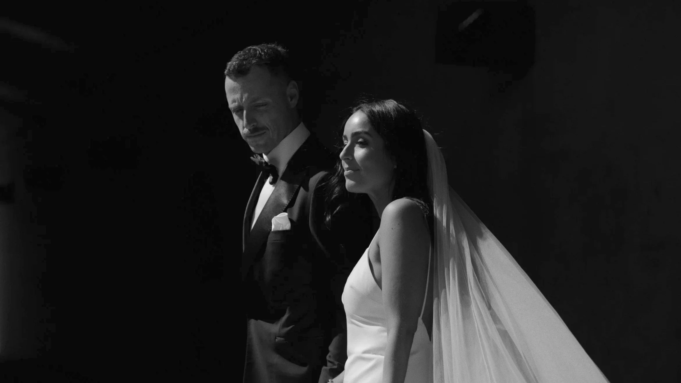 Black and white photo of a bride and groom in formal attire, standing side by side, with the groom looking down and the bride looking slightly to the side.
