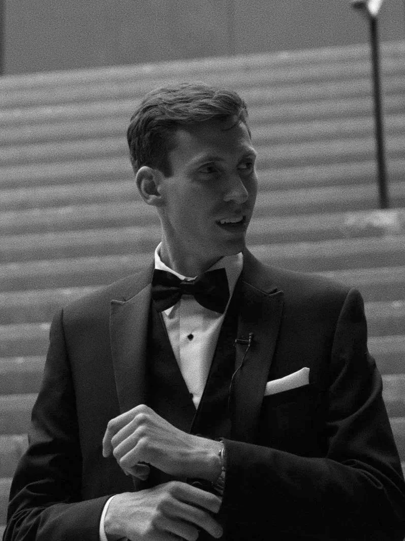 A young man dressed in a tuxedo with a bowtie, standing outdoors near stairs in downtown Ottawa, smiling.