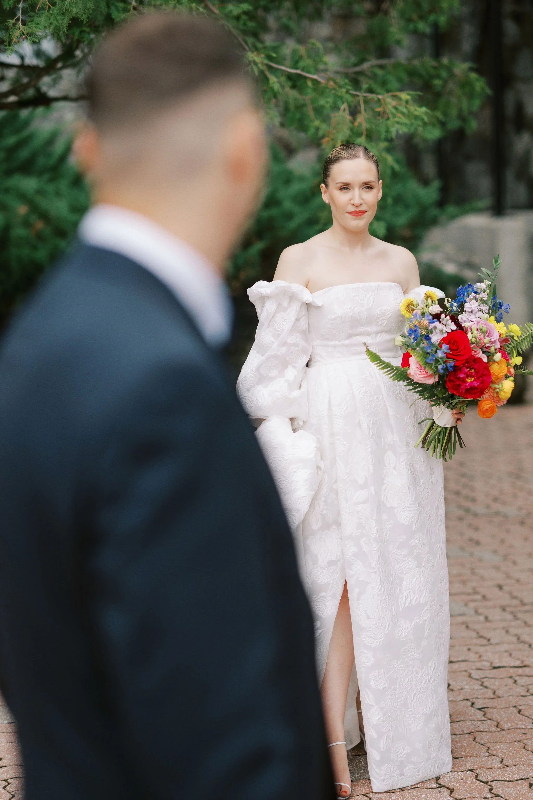 A woman in a white off-shoulder lace wedding dress holding a colorful bouquet of flowers, standing outdoors with green trees and a stone or brick pavement, while a blurred man in a suit appears in the foreground.