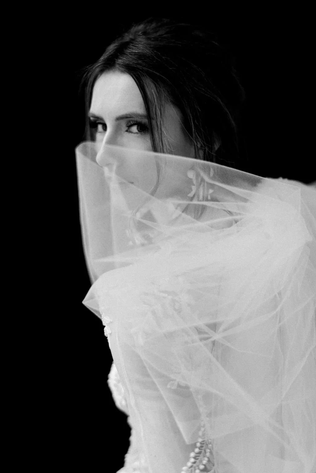 A black and white portrait of a woman with dark hair, partially obscured by sheer fabric, against a black background.