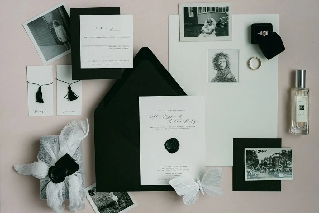 Flat lay of wedding invitation suite with black and white photos, gold ring, ring box, perfume, and decorative items on a light pink background.