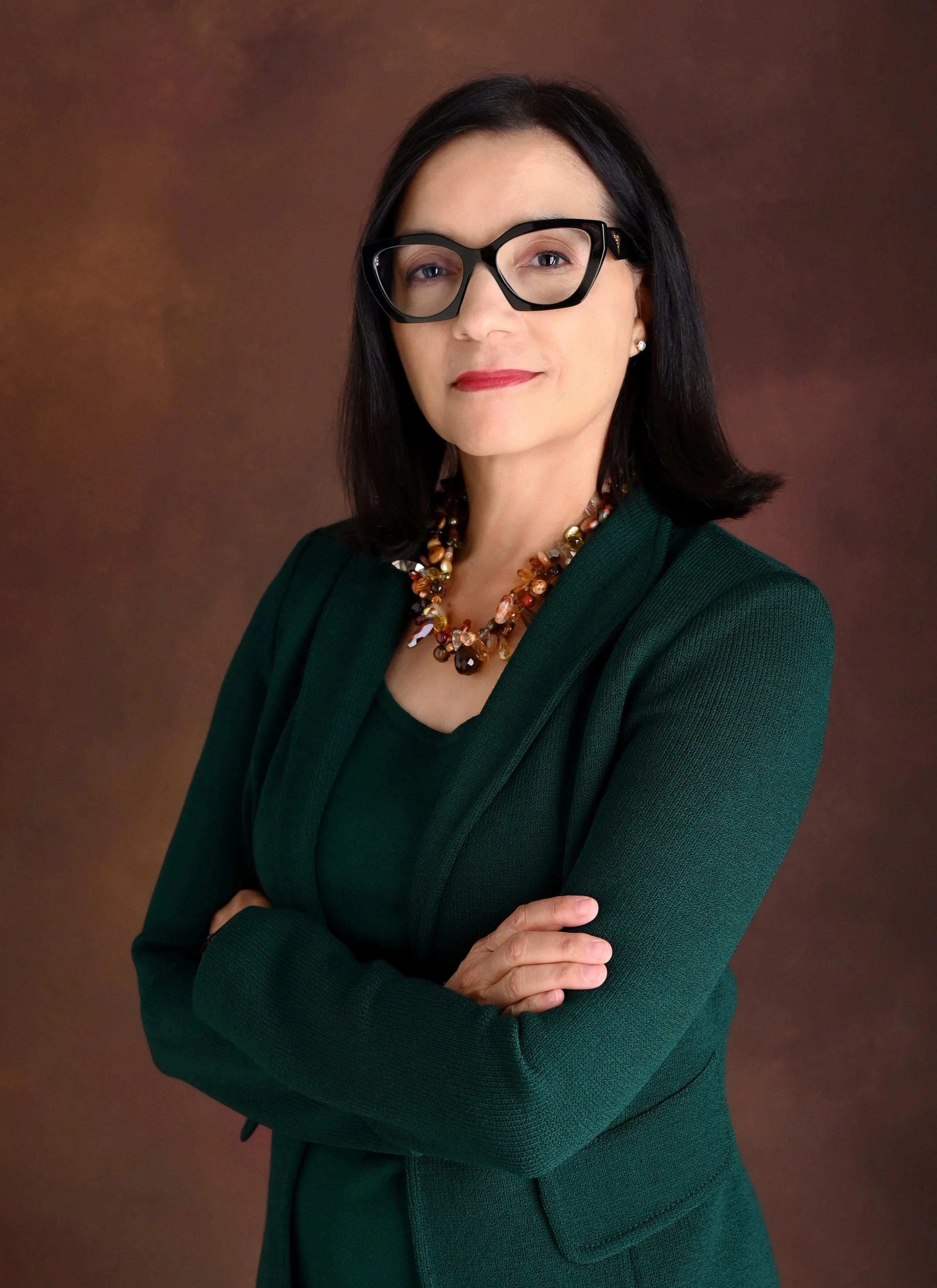 A professional woman with black hair, wearing glasses, a dark green blazer, and a colorful beaded necklace, standing with her arms crossed against a brown background.