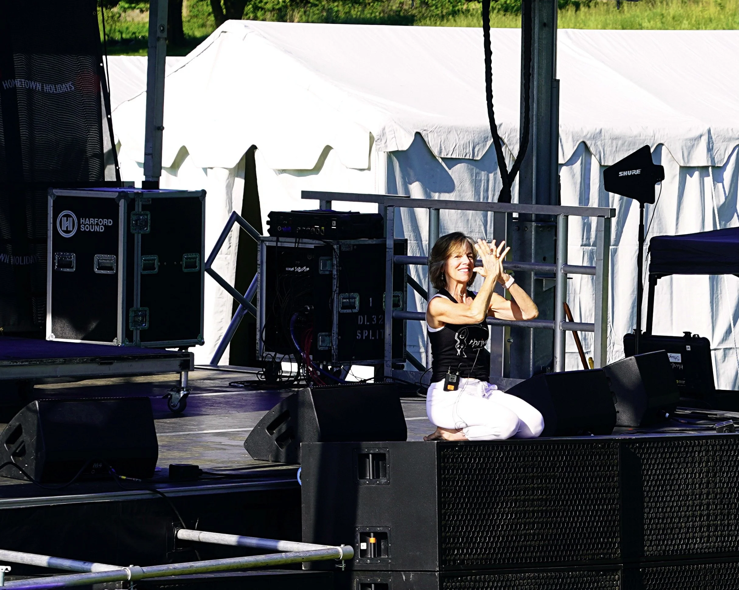 A woman kneeling on stage with a microphone headset, smiling and gesturing with her hands, surrounded by sound equipment and speakers, with a white tent and green foliage in the background.