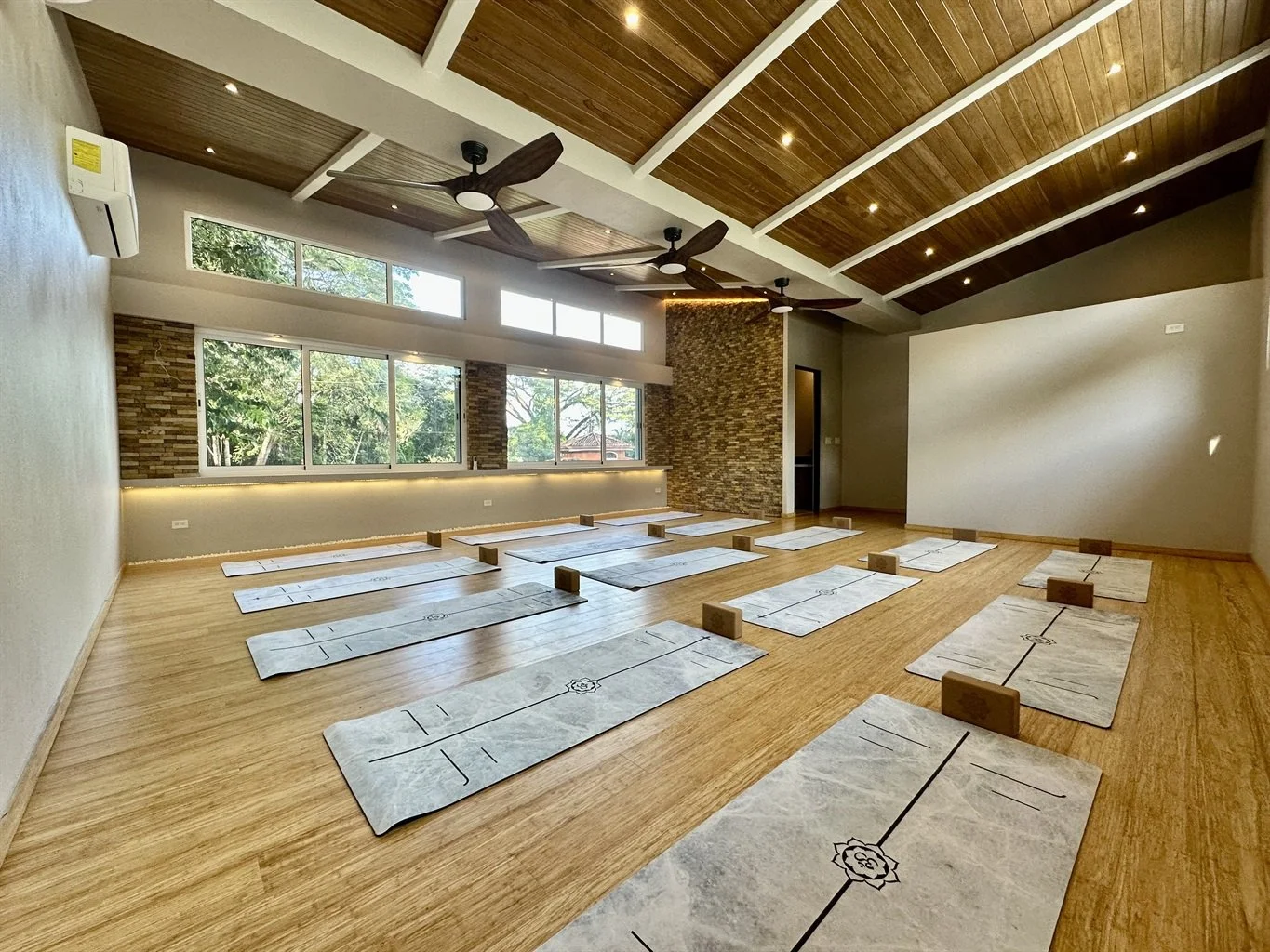 Indoor yoga or meditation class setup with mats and blocks on a wooden floor, large windows, wood-paneled ceiling with fans, and a brick accent wall.