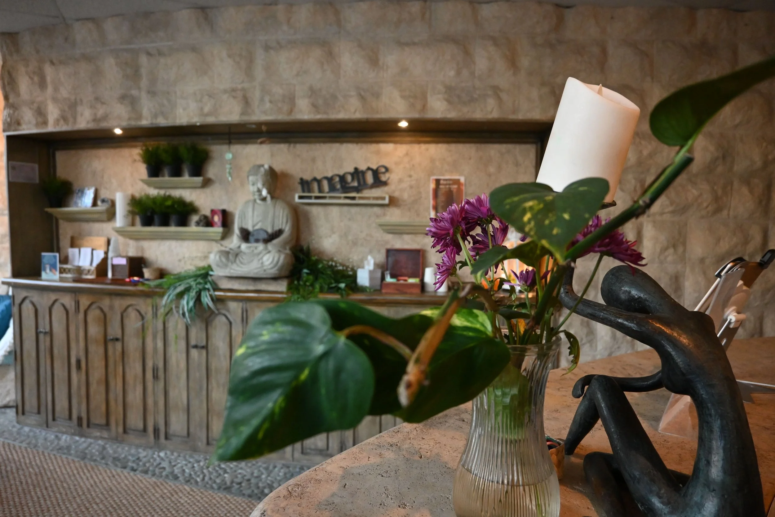 A glass vase with purple flowers and green leaves on a table in the foreground, with a stone wall and decorative decor including a statue, shelves with potted plants, a sign that says 'imagine', and framed pictures in the background.