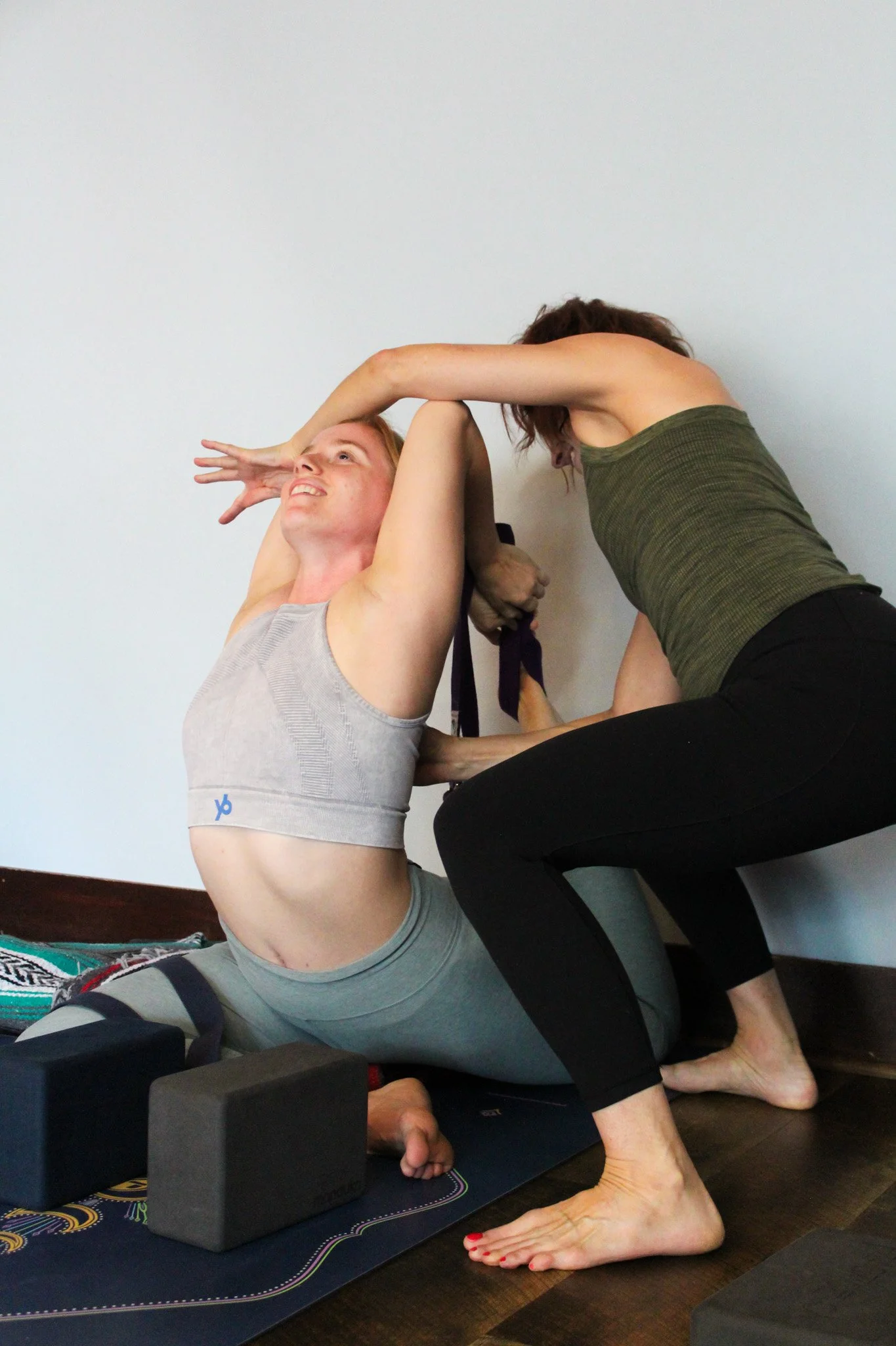 Two women practicing partner yoga in a room with a plain white wall. One woman is sitting on the floor with her legs bent and arms elevated, while the other woman is leaning over her, grasping her arm and her waist, supporting her in a yoga pose.