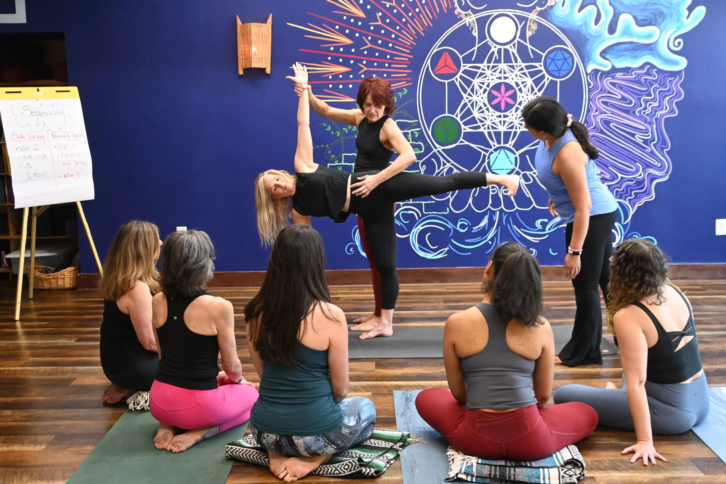 A woman demonstrates a yoga pose on a black mat in front of an audience of seated women in a yoga class. The background features a vibrant blue wall with cosmic and geometric artwork.