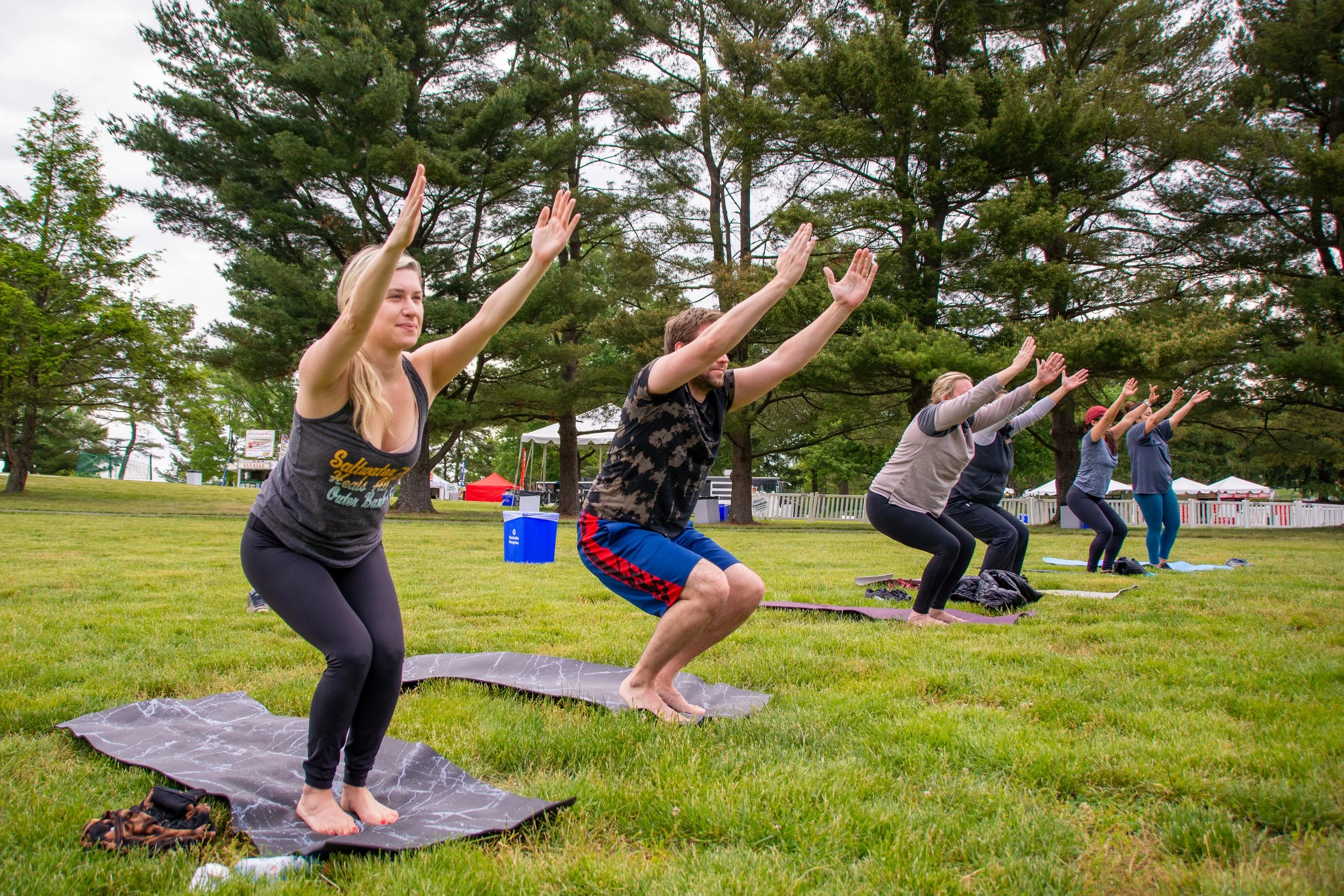 A group of people practicing yoga outdoors on a grassy lawn, with large trees and festival tents in the background.