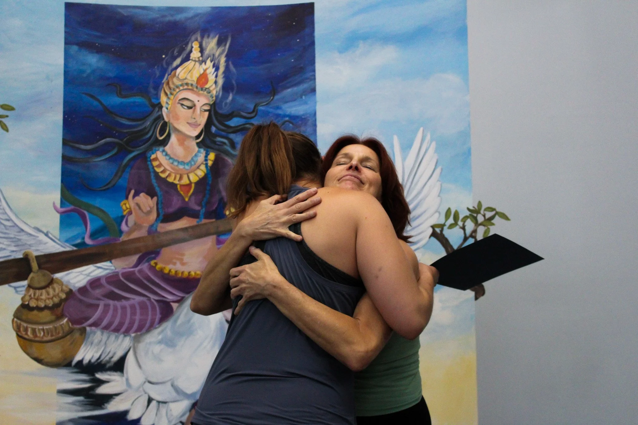 Two women hugging each other in front of a colorful mural of a Hindu goddess with dark hair and jewelry, set against a blue sky background with clouds and a small tree.