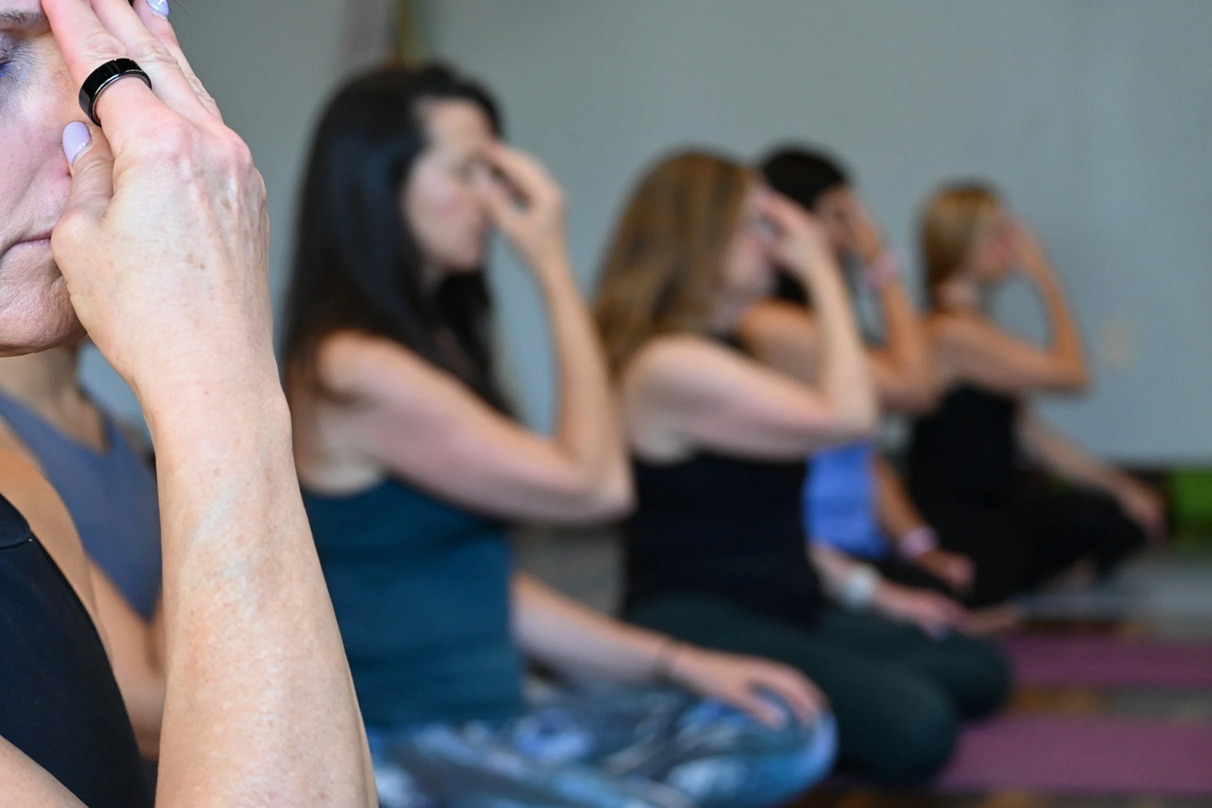 Several women sitting in a row, practicing meditation or mindfulness, with one woman in the foreground pressing her hands together in a prayer position.
