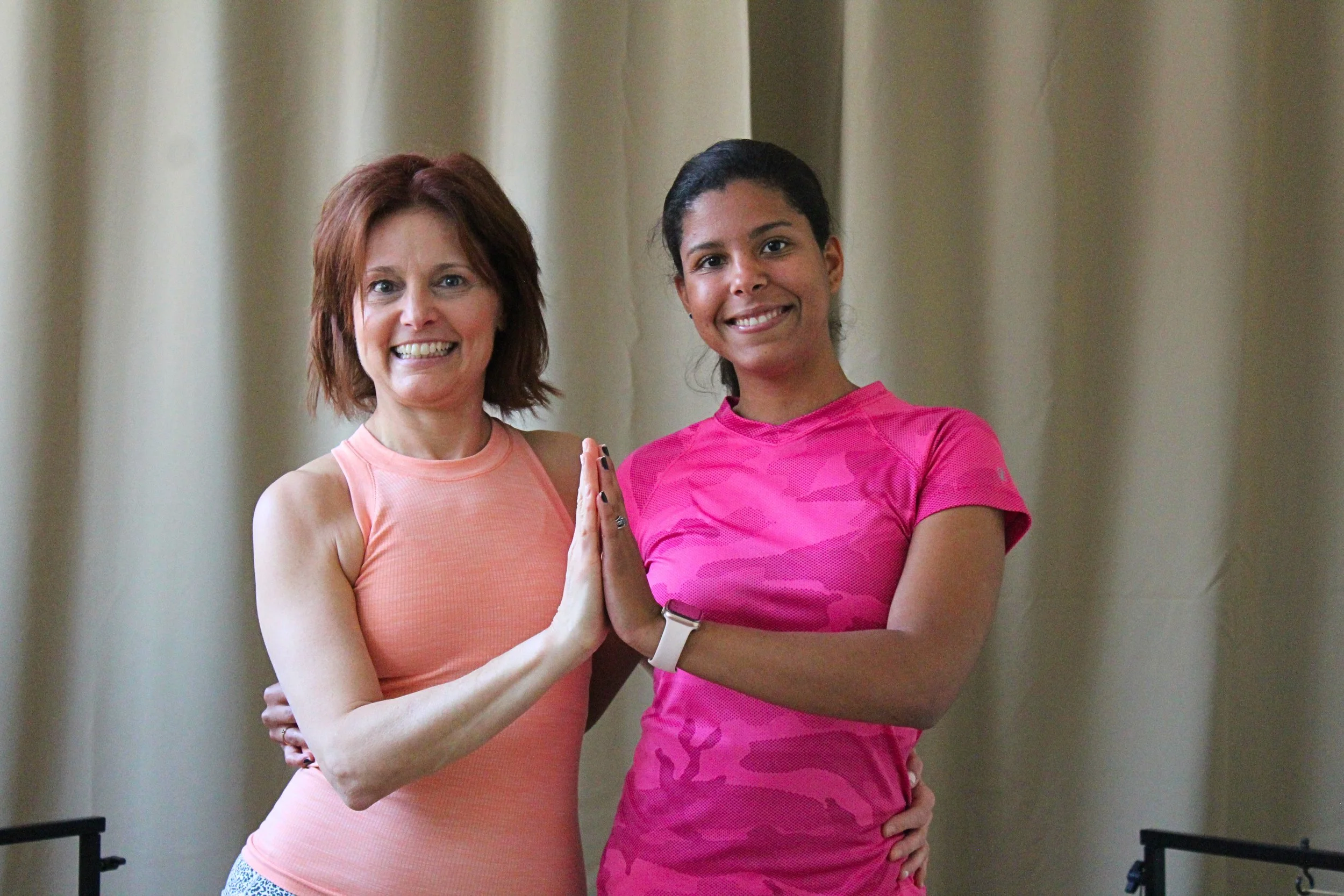 Two women, one with red hair and one with black hair, smiling and giving each other a high five in fitness gear, in a room with beige curtains.