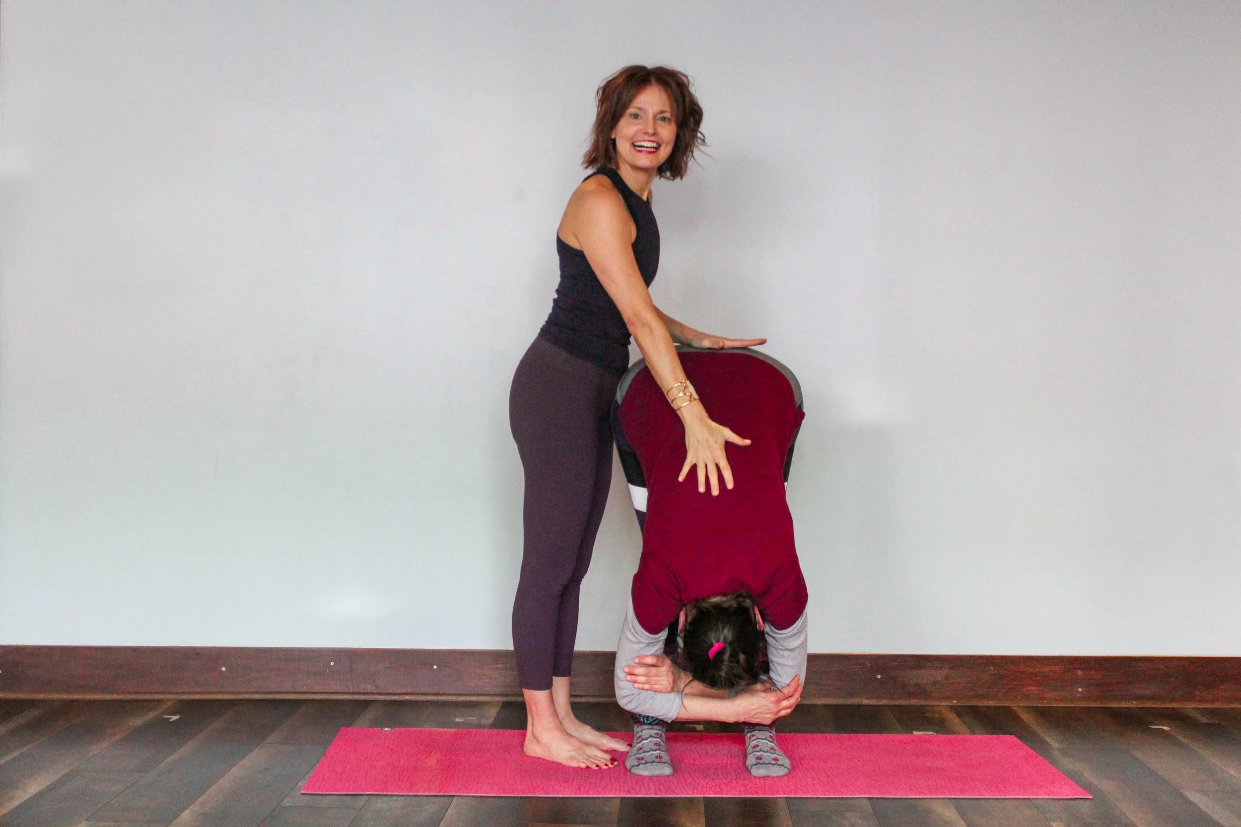 A woman practicing yoga in a headstand position with another woman supporting her by holding her legs, both on a yoga mat in front of a plain white wall.