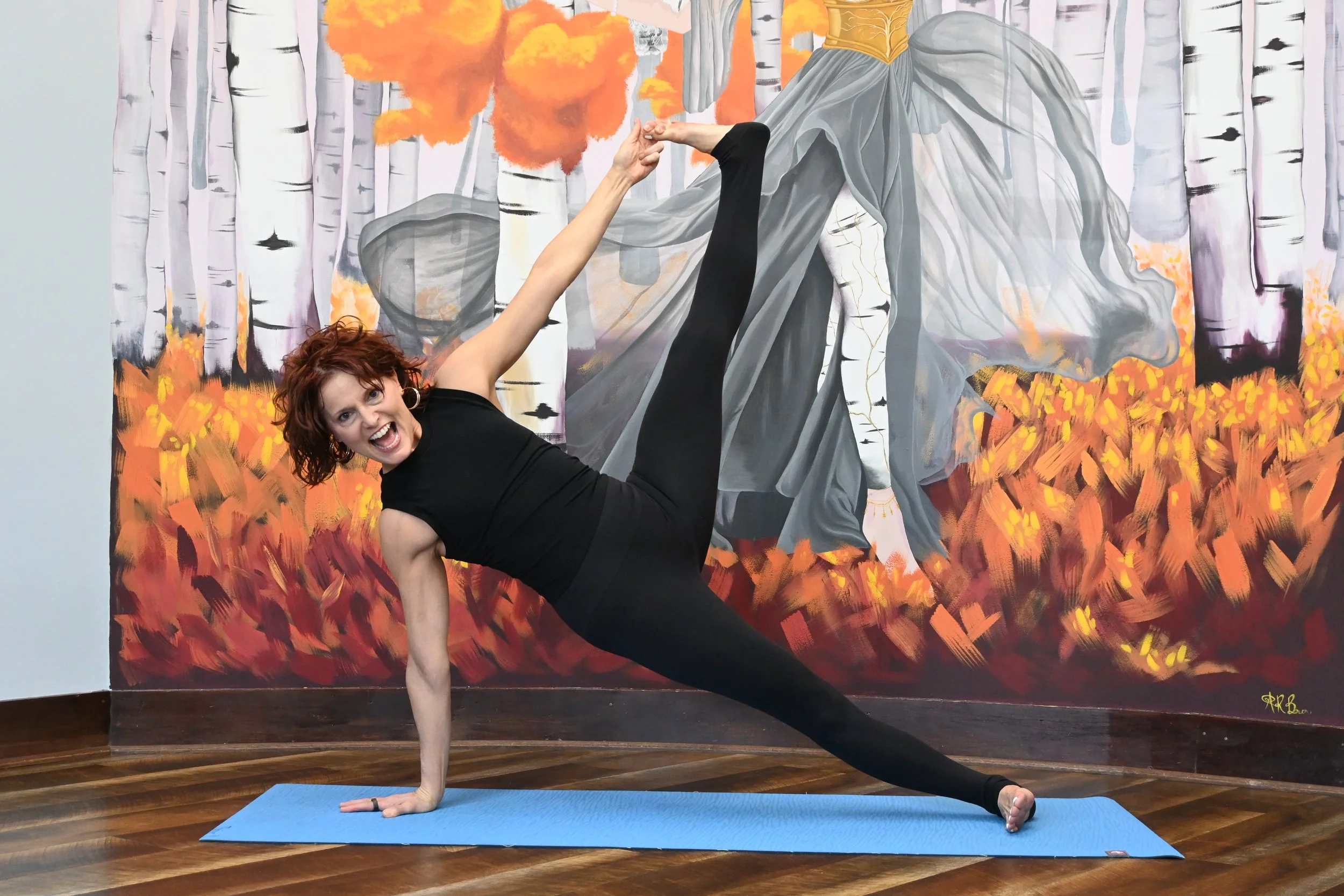 A woman with curly red hair performing a yoga pose on a blue mat in front of a large painting of a woman with gray hair, standing on a forest path with autumn trees, while holding her foot above her head with one hand.