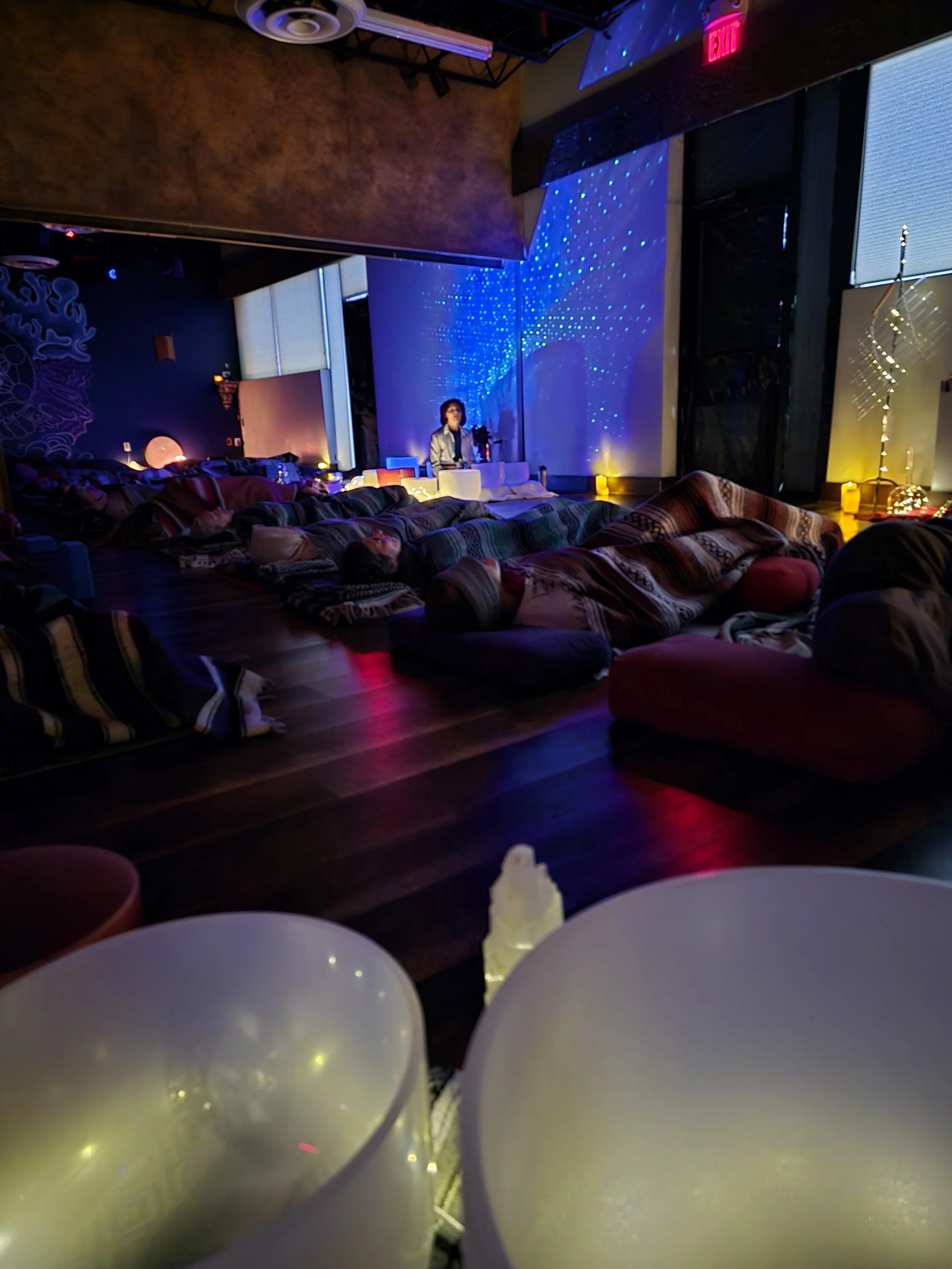 Indoor space with purple and blue lighting, cushions on the floor, a person sitting at a table, and a wall decorated with blue fairy lights. There are white balloons in the foreground and candles along the wall, creating a cozy ambiance.