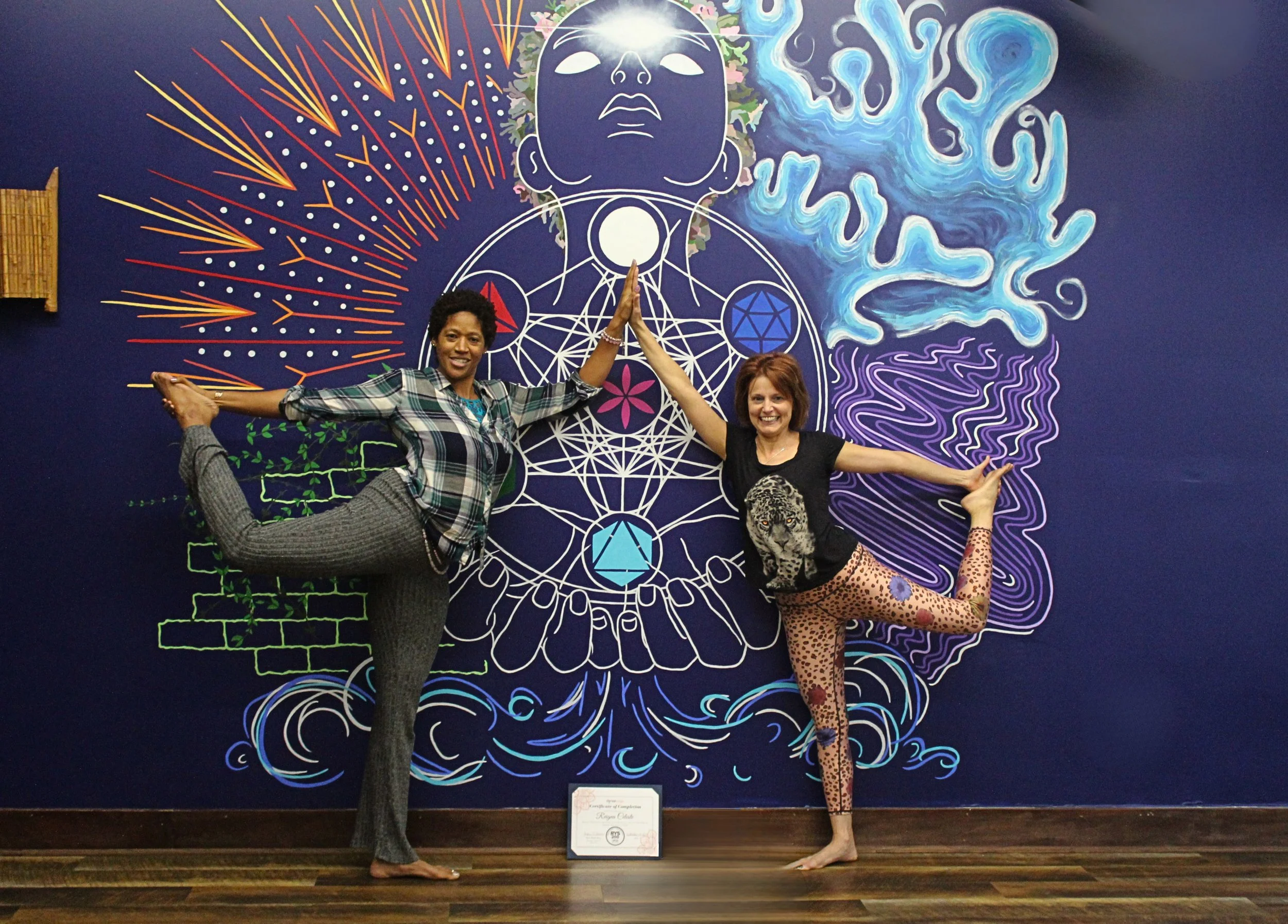 Two women pose in front of a colorful, abstract mural featuring geometric and organic shapes, with one woman balancing on one leg and the other with a leopard print outfit.