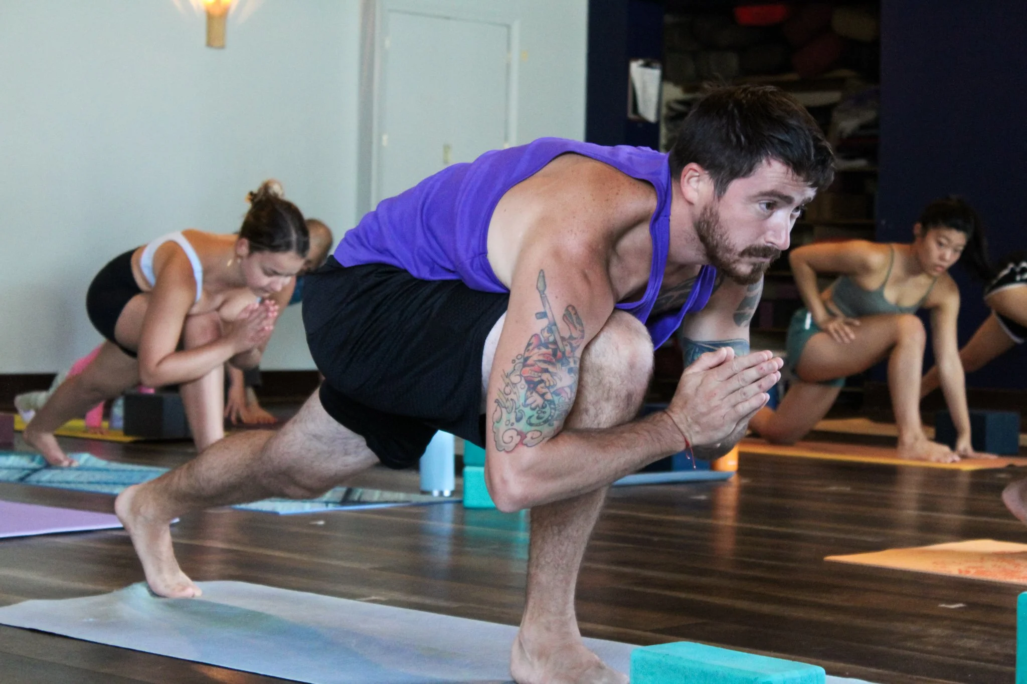 A man with tattoos on his arms practicing yoga in a yoga studio with others, all in the plank pose, balancing on one arm and one leg, on yoga mats.