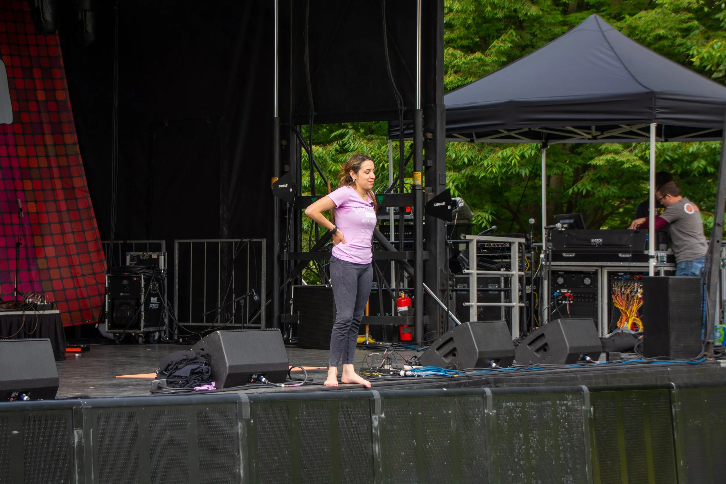 A woman on an outdoor stage with musical equipment, standing barefoot with her arms akimbo, wearing a purple t-shirt and gray pants, surrounded by trees, with a person adjusting sound equipment in the background.
