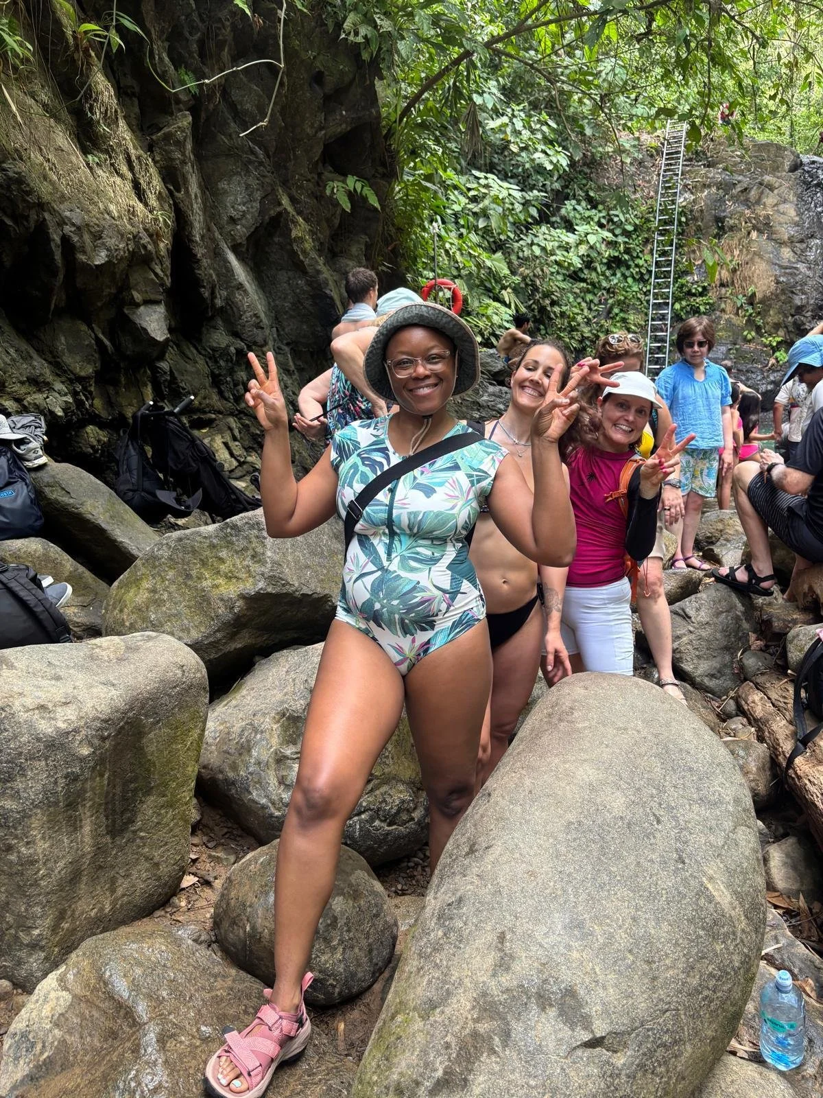 Group of women and men on a rocky trail in a lush, green forest, some posing with peace signs, among climbing gear and backpacks.