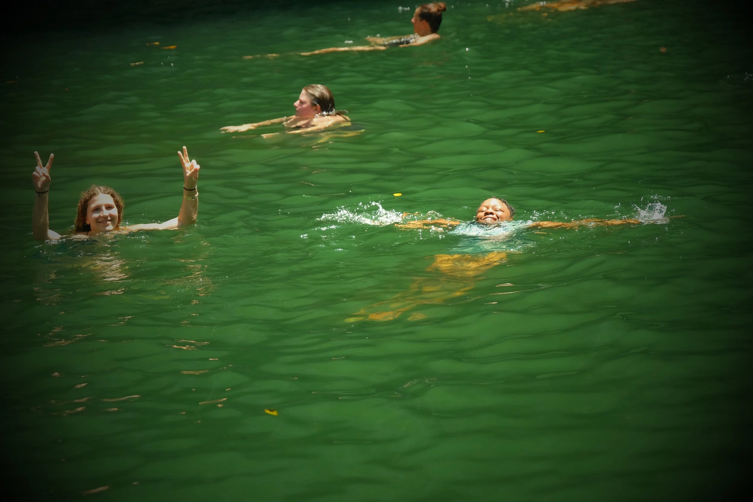 Four women swimming and floating in a green-tinted natural water body, with one woman smiling and giving a peace sign, others relaxing and swimming.
