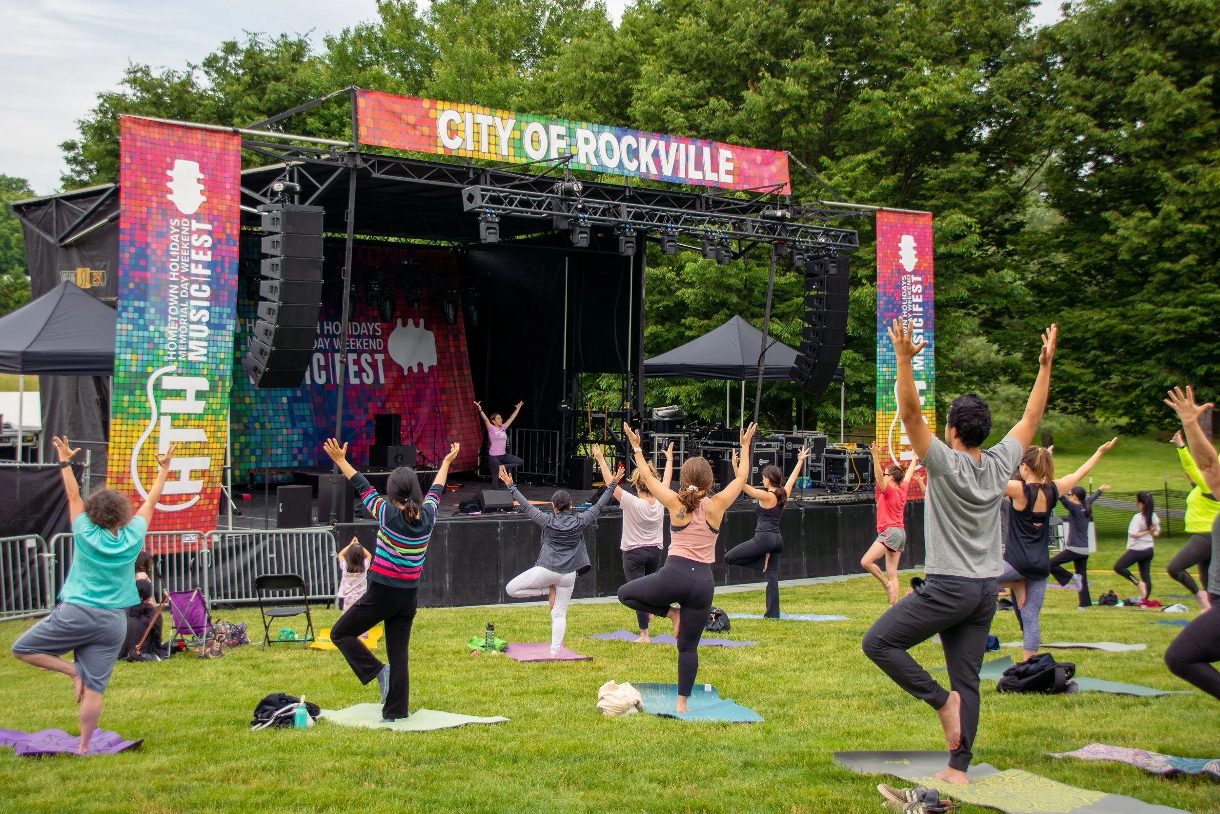 People participating in an outdoor yoga class on a grassy area with an event stage in the background during daytime.